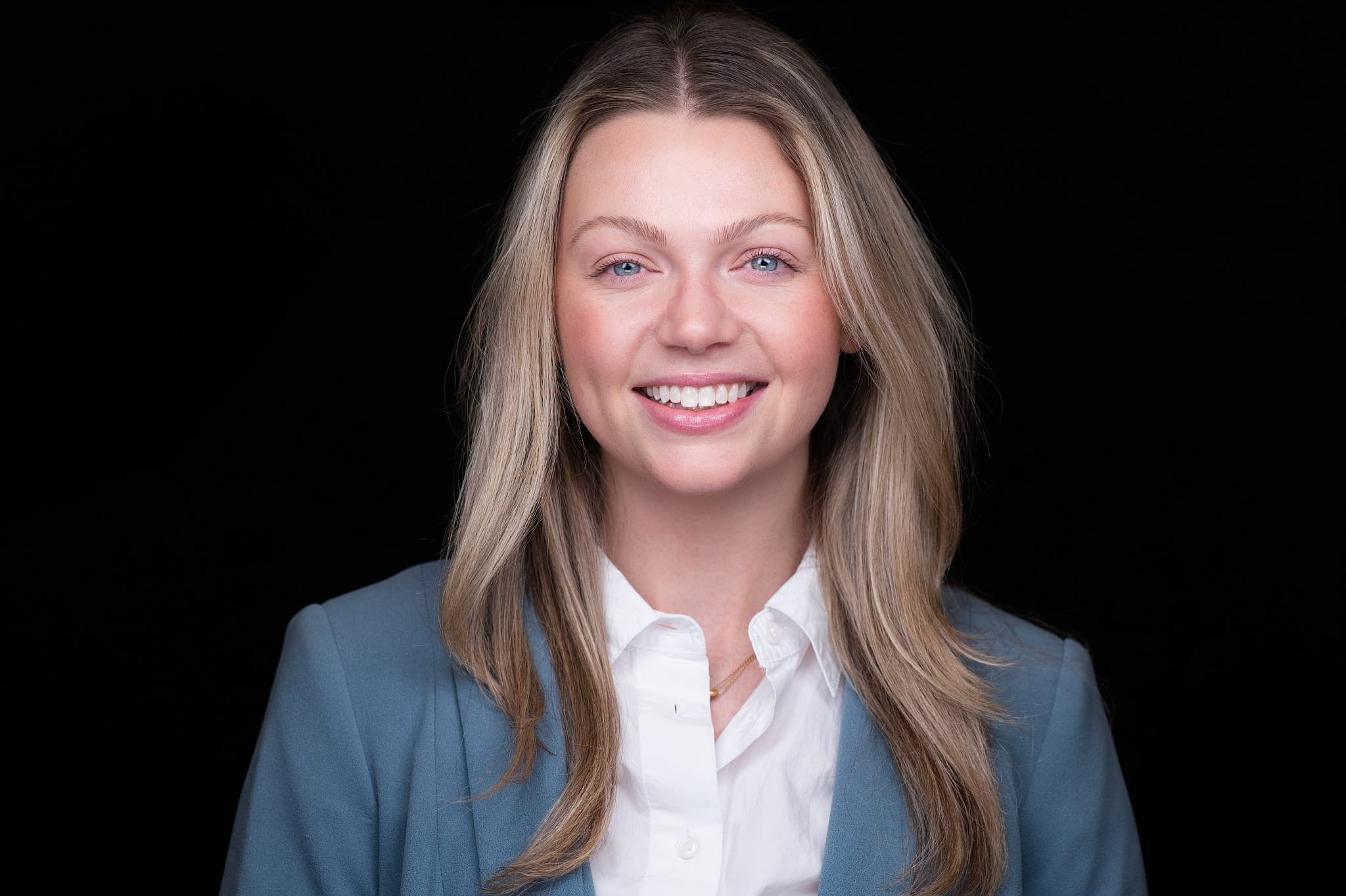 Professional corporate headshot of woman in blue blazer and white shirt with warm smile against black studio background