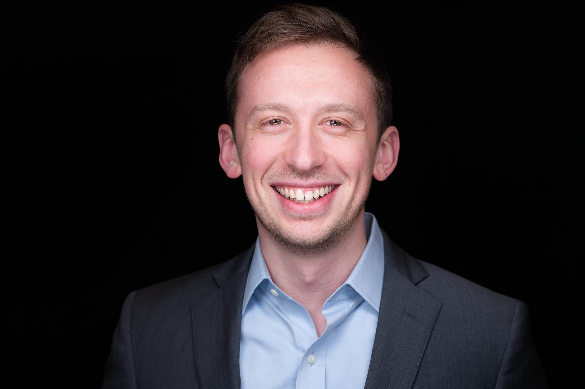 Executive headshot of man in navy blazer and light blue shirt with genuine smile against black studio background