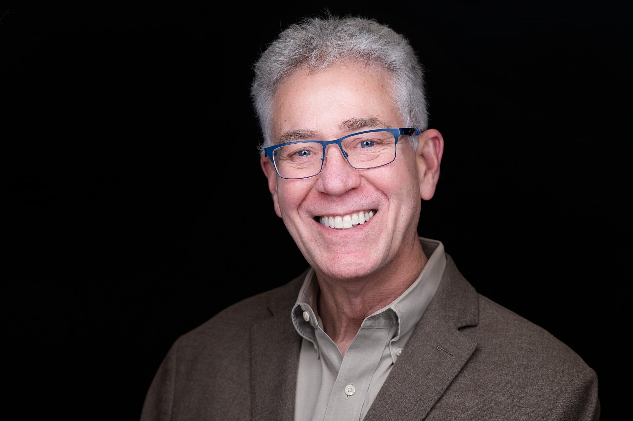 Mature professional headshot of man with grey hair, glasses, and brown blazer with confident friendly expression