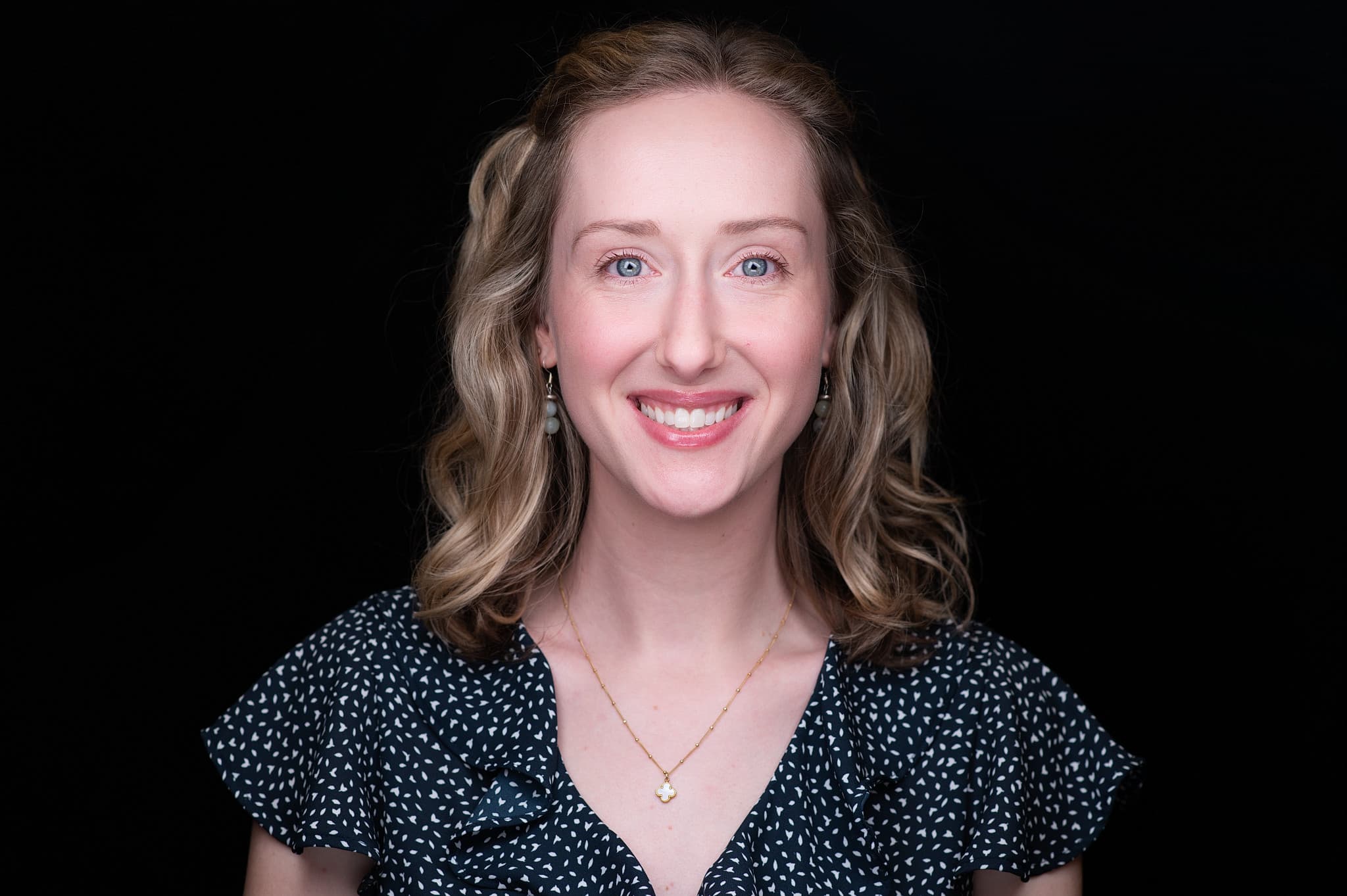 Professional headshot of woman with shoulder-length blonde hair, navy patterned top, and genuine smile against black background