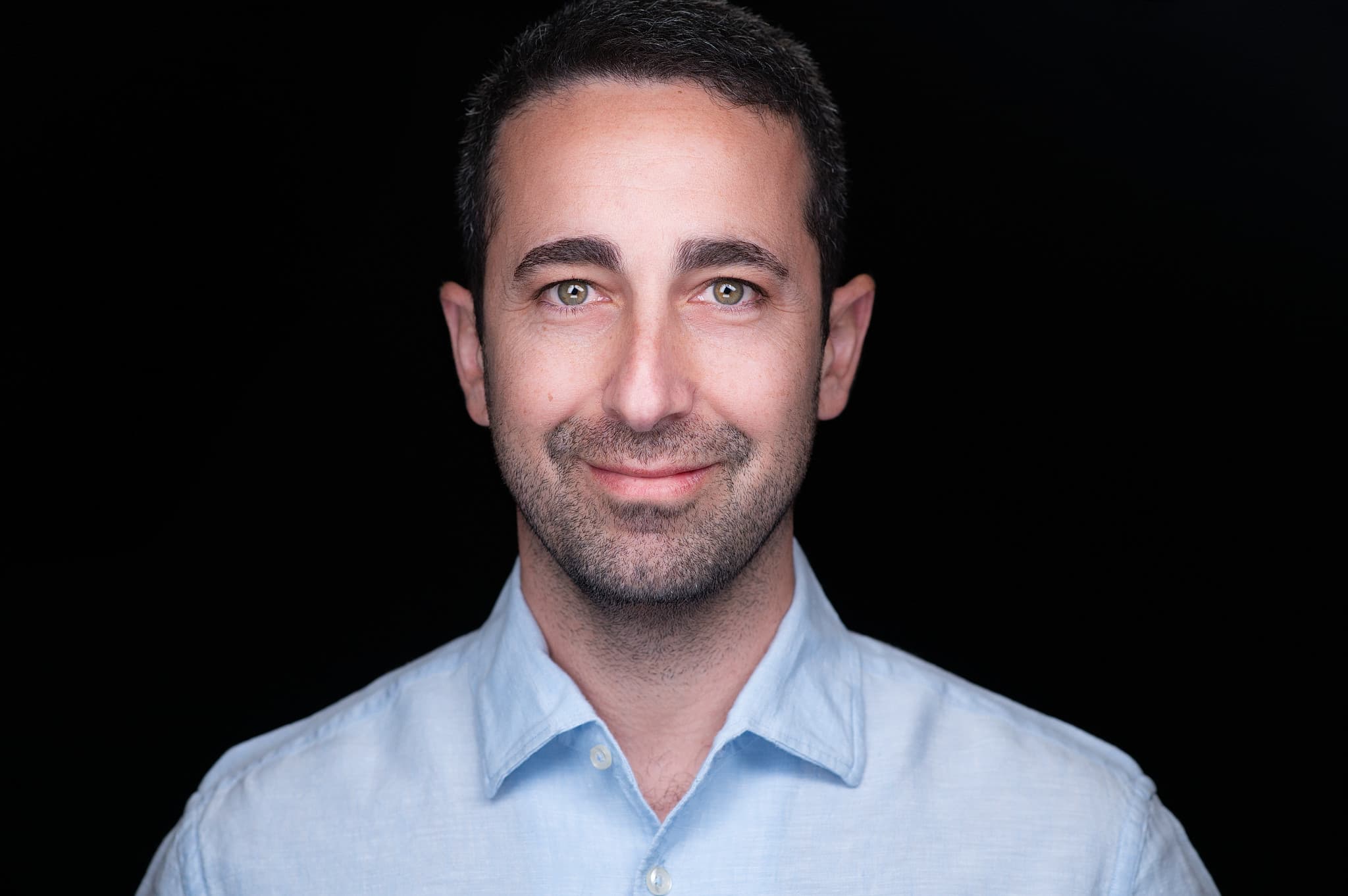 Confident male professional headshot in light blue shirt against black background with studio lighting