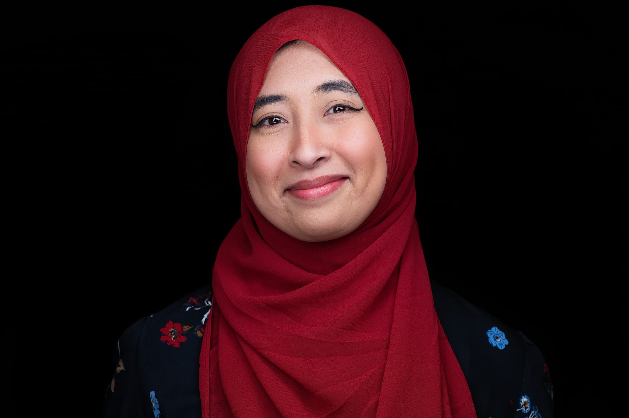 Warm professional headshot of female wearing red hijab and navy embroidered garment against black background