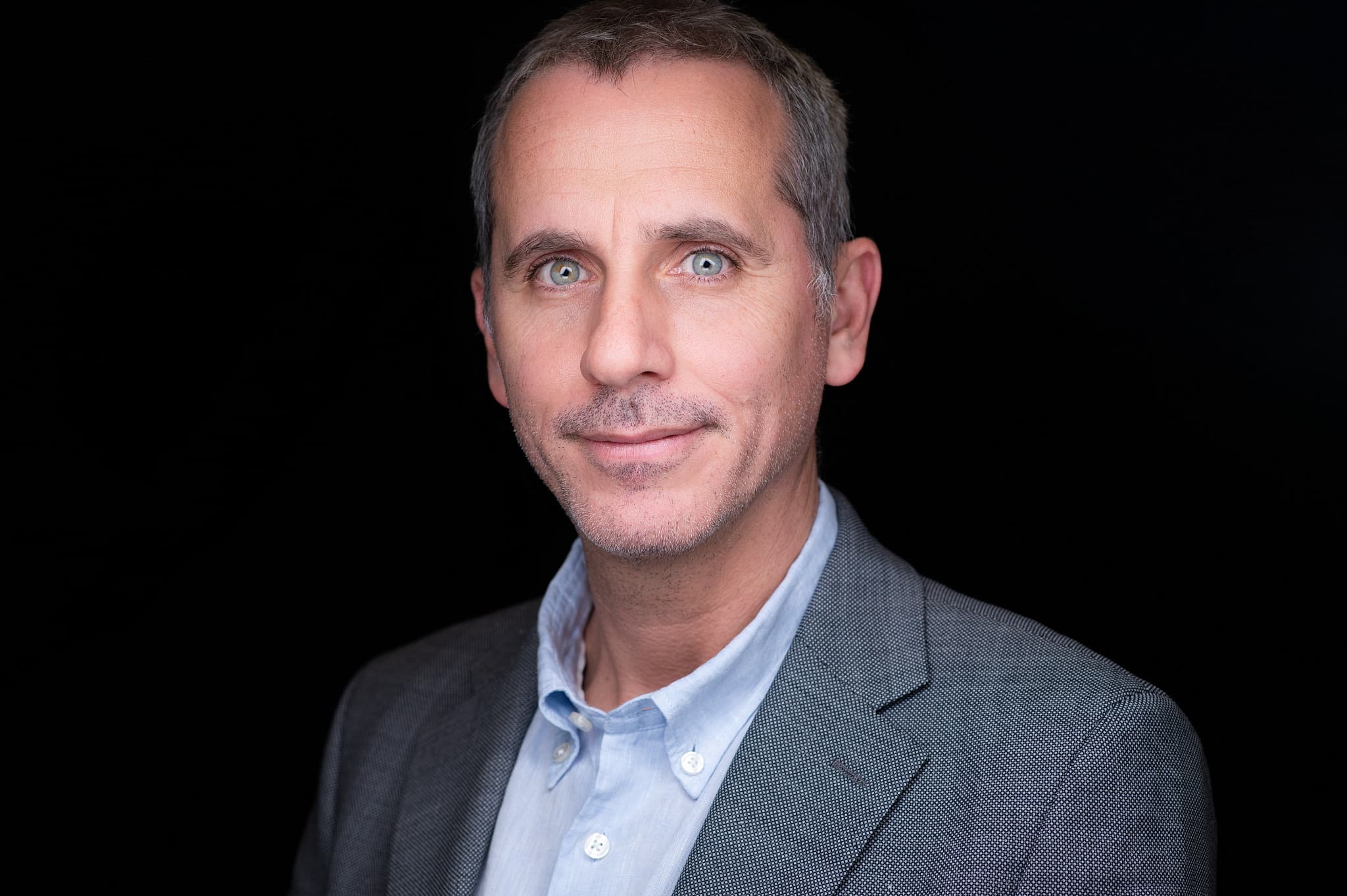Composed male professional headshot in blue shirt and gray blazer with direct gaze and calm expression