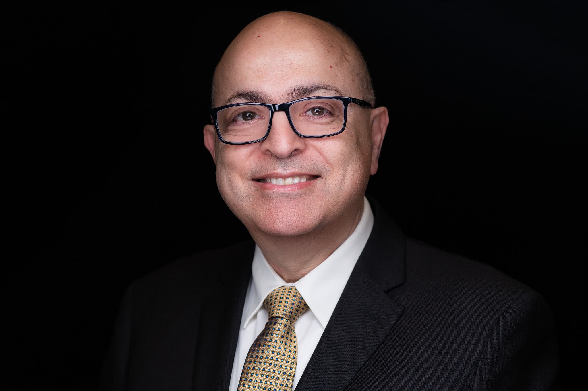 Personable male professional headshot wearing glasses, navy suit, patterned gold and blue tie