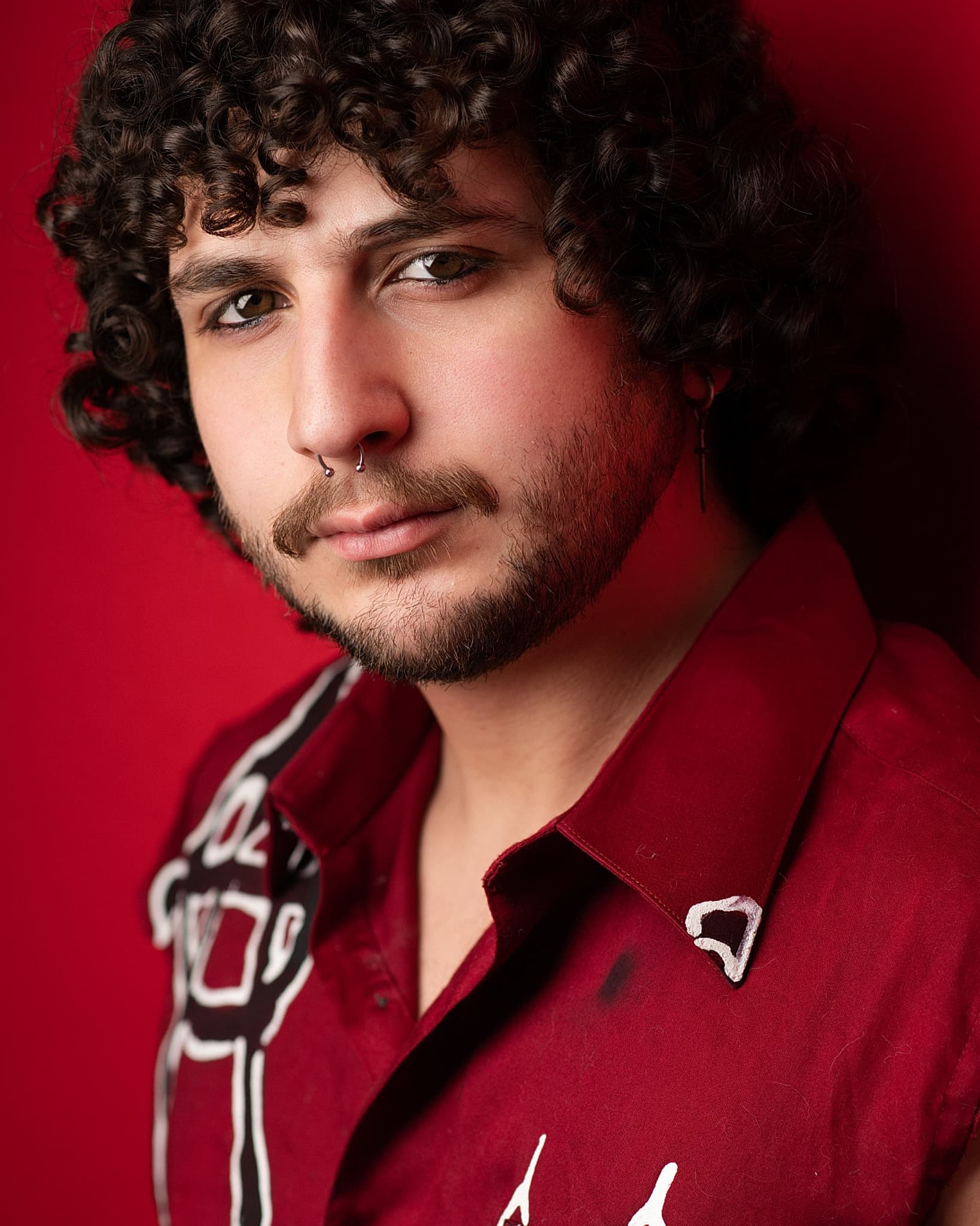 Atlanta actor headshot of Aiden Anzaldúa, young man with dark curls, a mustache and a nose ring in a deep red shirt against a red studio backdrop