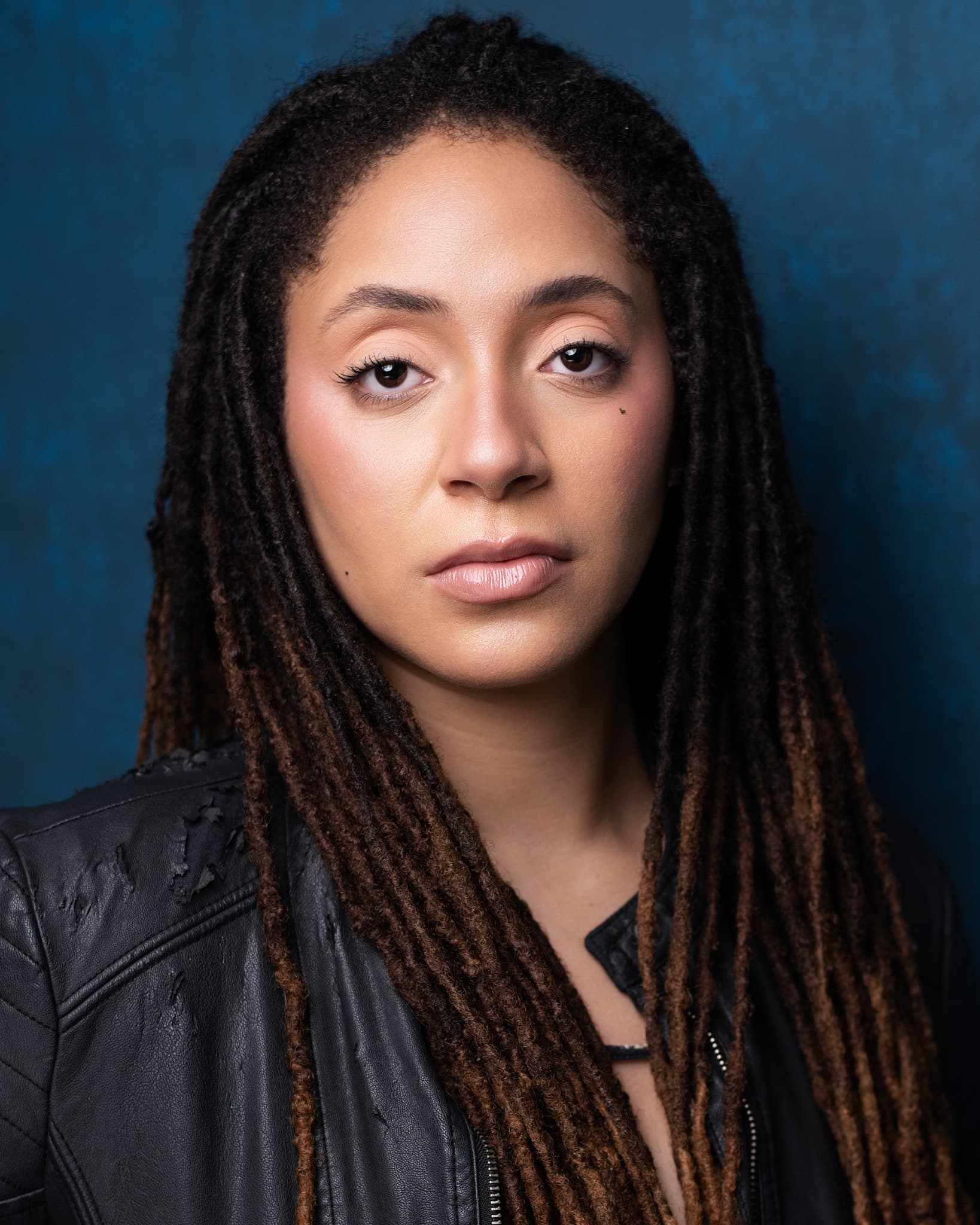 Atlanta theatrical actor headshot of Ajeya Brandon, woman with long ombre locs in a black leather jacket with an intense gaze against a moody teal-blue backdrop