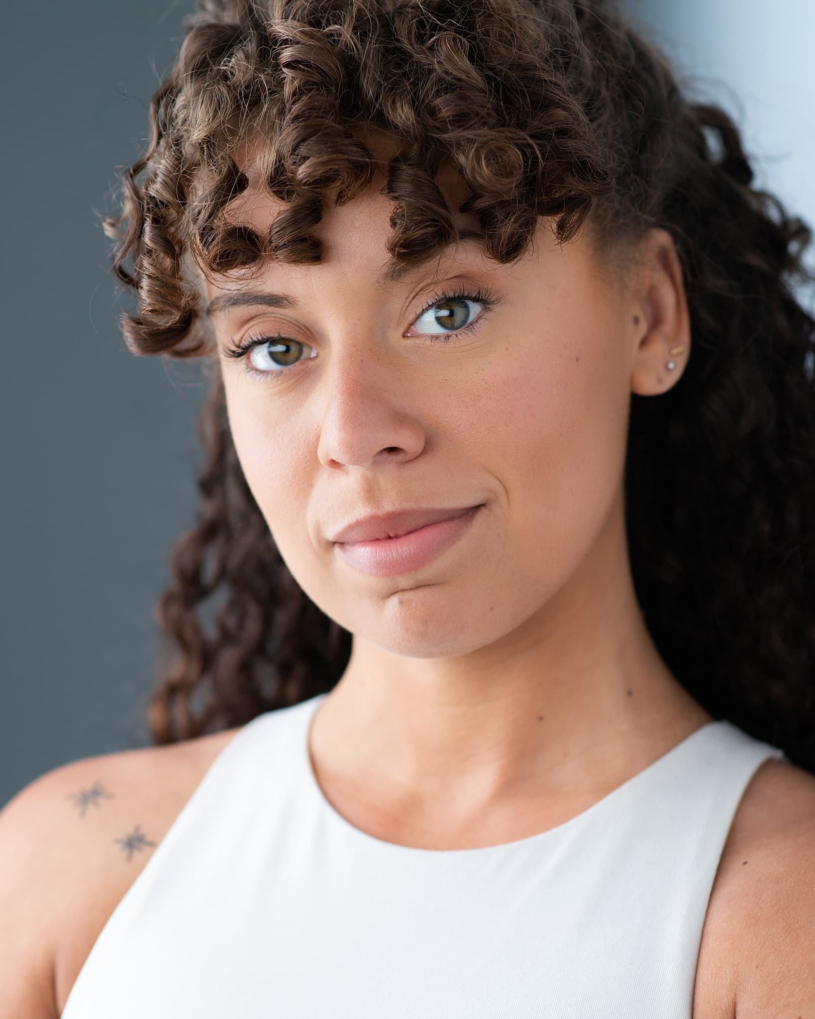 Atlanta commercial actor headshot of Chelsey Goldsmith, woman with curly brown hair and green eyes in a white tank top with a subtle smile against a gray background