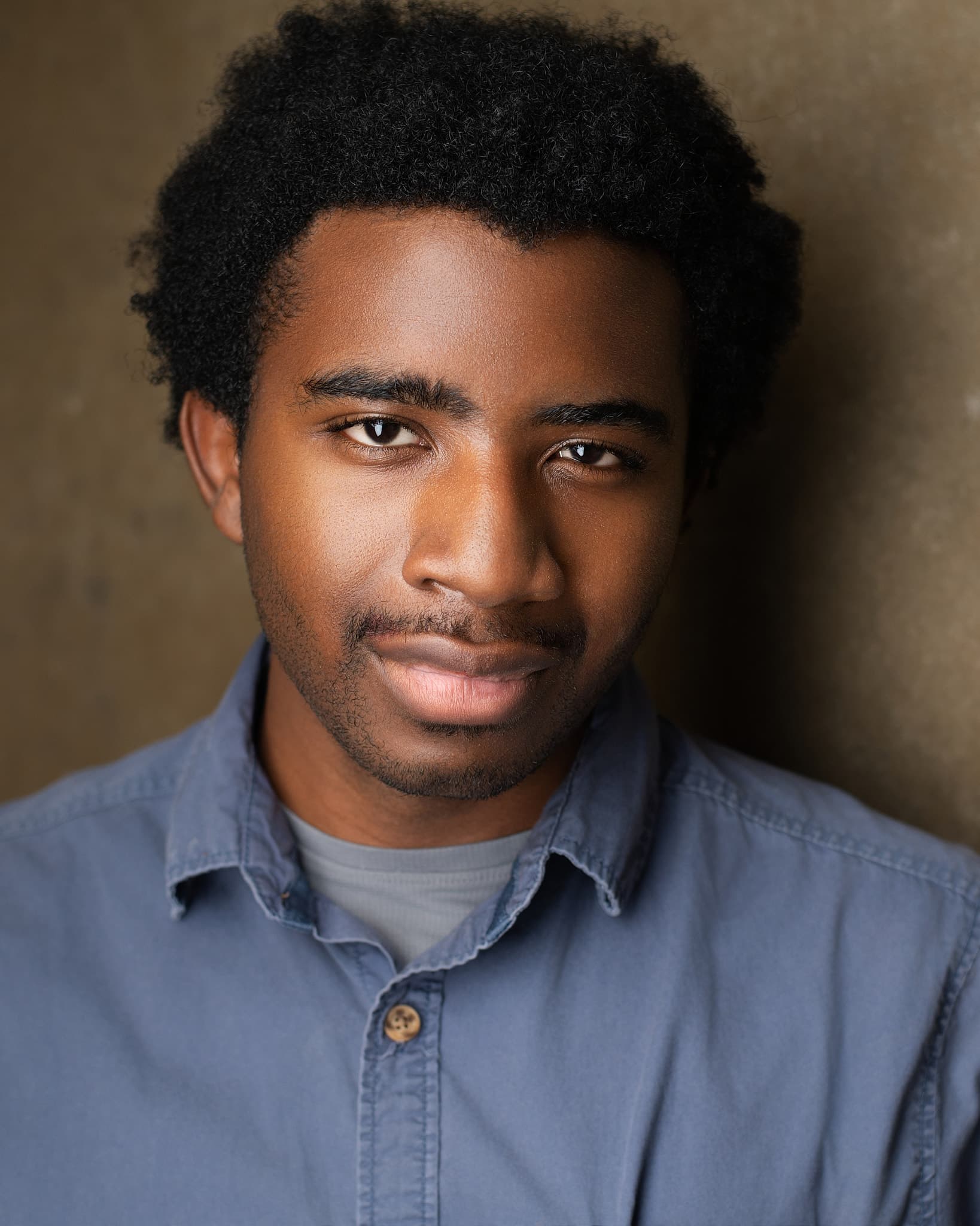 Atlanta actor headshot of Christopher Cox, young man with an afro in a blue chambray shirt with a calm expression against a warm tan backdrop