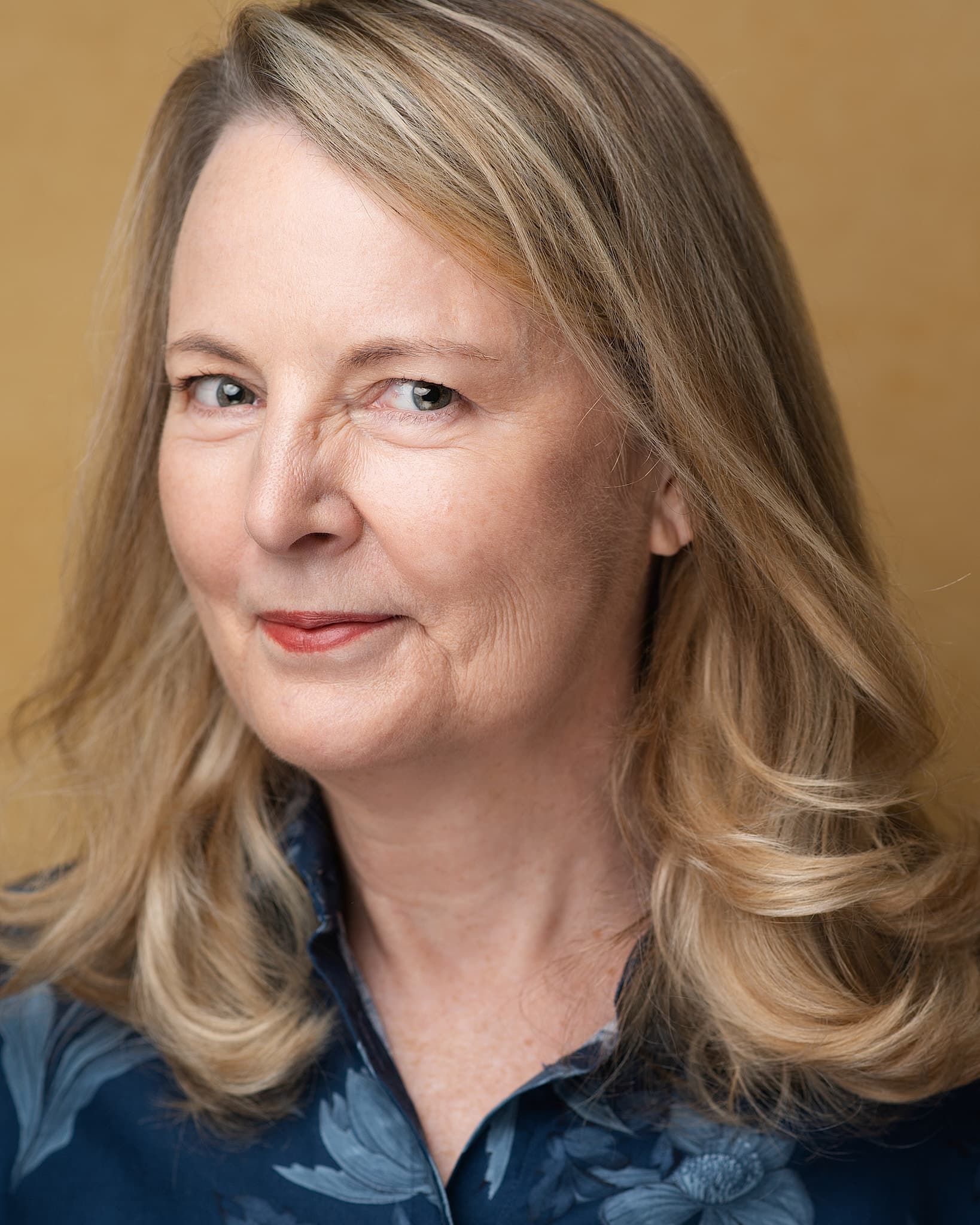 Atlanta theatrical actor headshot of Elisa Carlson, mature blonde woman in a blue floral blouse looking off camera against a warm gold backdrop