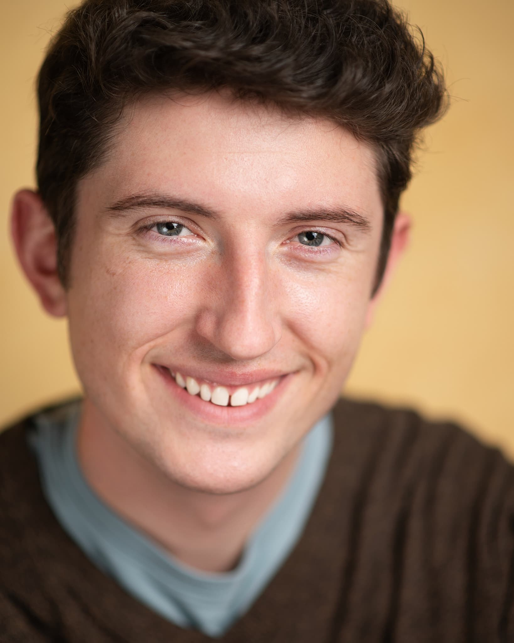 Atlanta commercial actor headshot of Jobe Kerr, young man with dark curly hair and a bright smile in a brown sweater against a warm cream backdrop