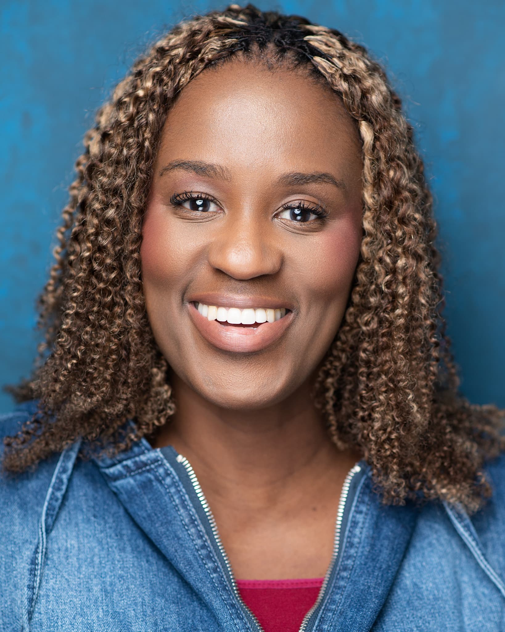 Atlanta commercial actor headshot of Myiesha Freeman, woman with curly honey-highlighted hair and a bright smile in a denim jacket against a blue backdrop