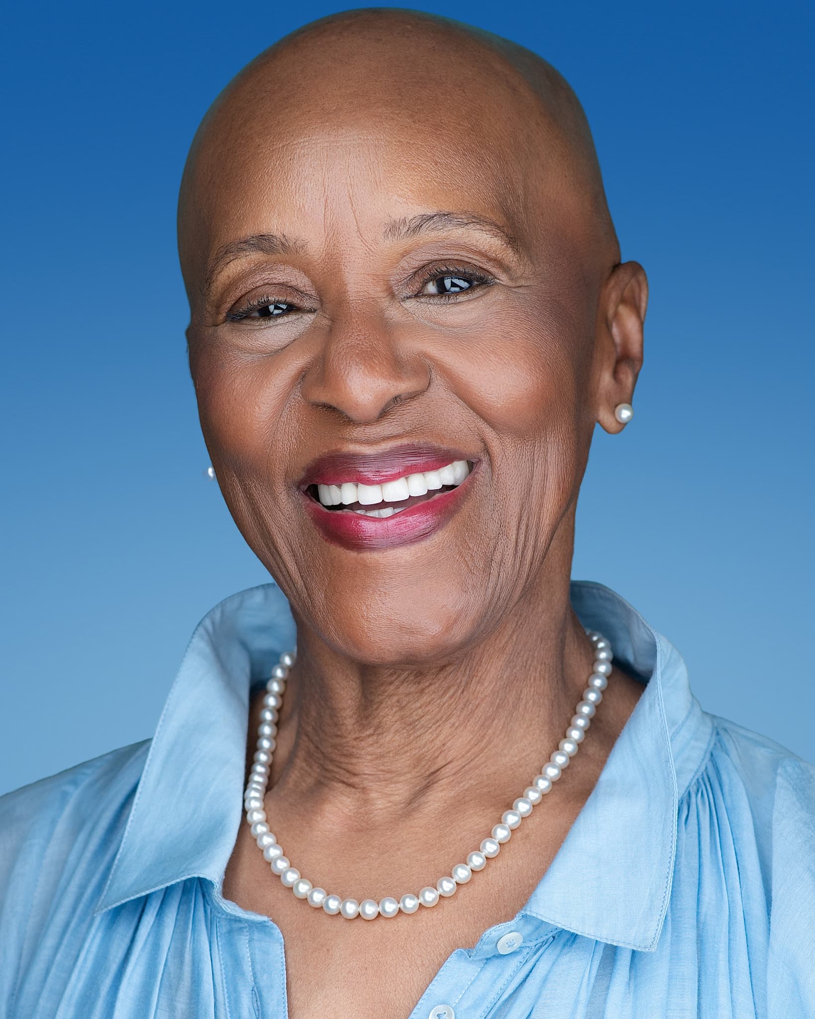 Atlanta commercial actor headshot of Pamela Garvey, mature woman with a shaved head and a radiant smile wearing pearls and a light blue blouse against a blue backdrop