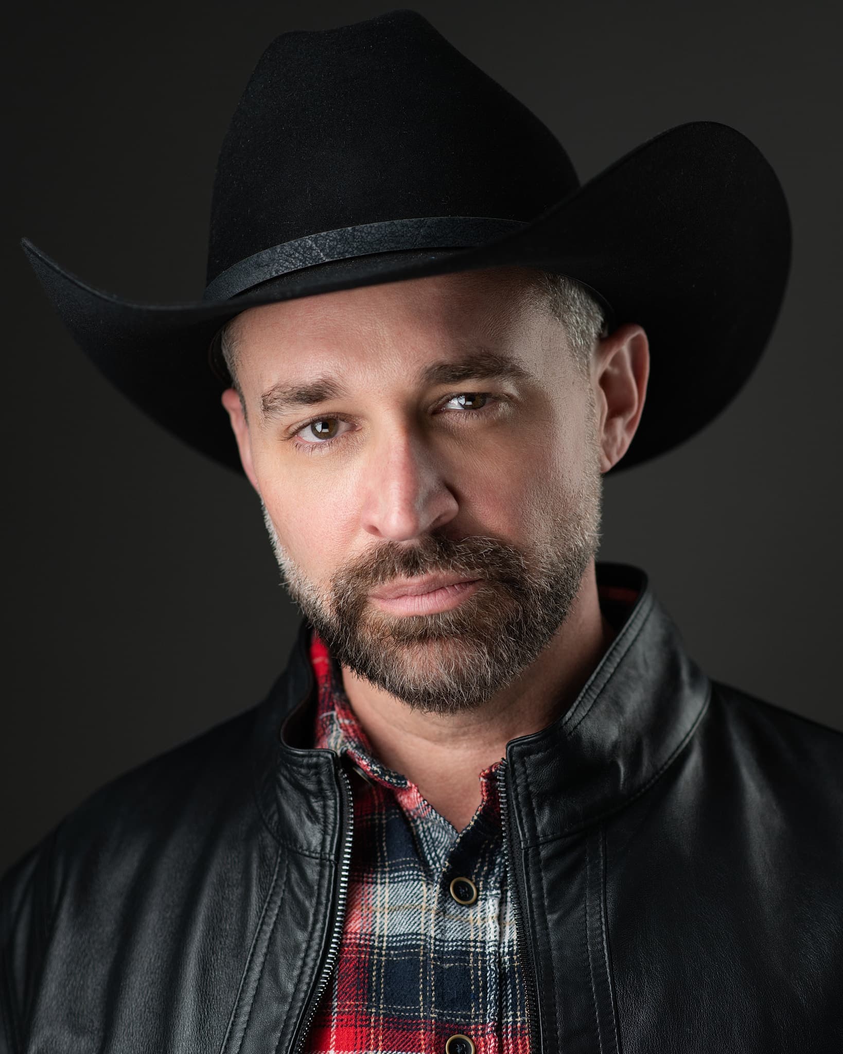 Atlanta theatrical actor headshot of Thad Garland, bearded man in a black cowboy hat and leather jacket over red plaid against a dark studio background