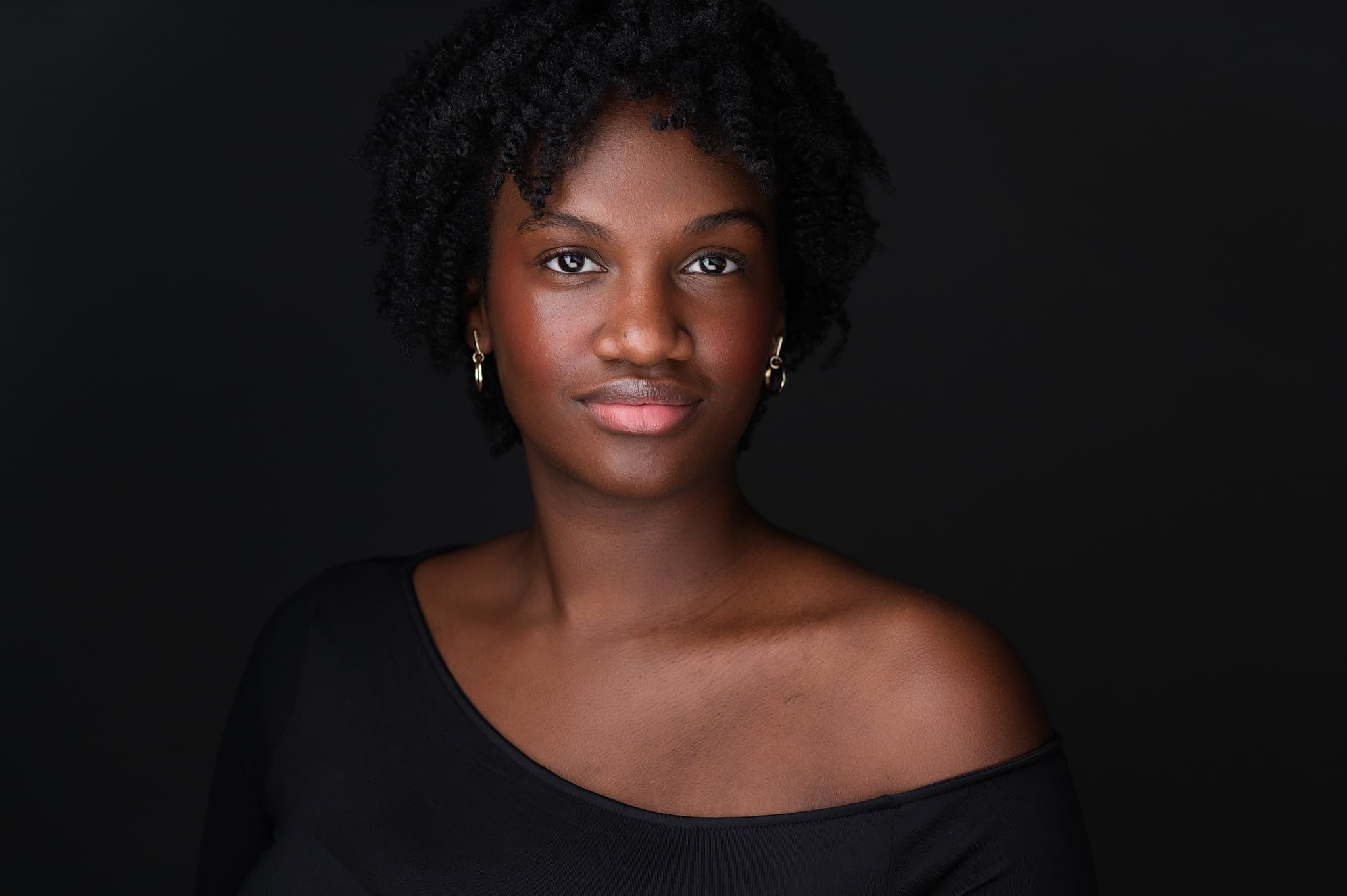 Theatrical headshot of young actress in black off-shoulder top with dark backdrop