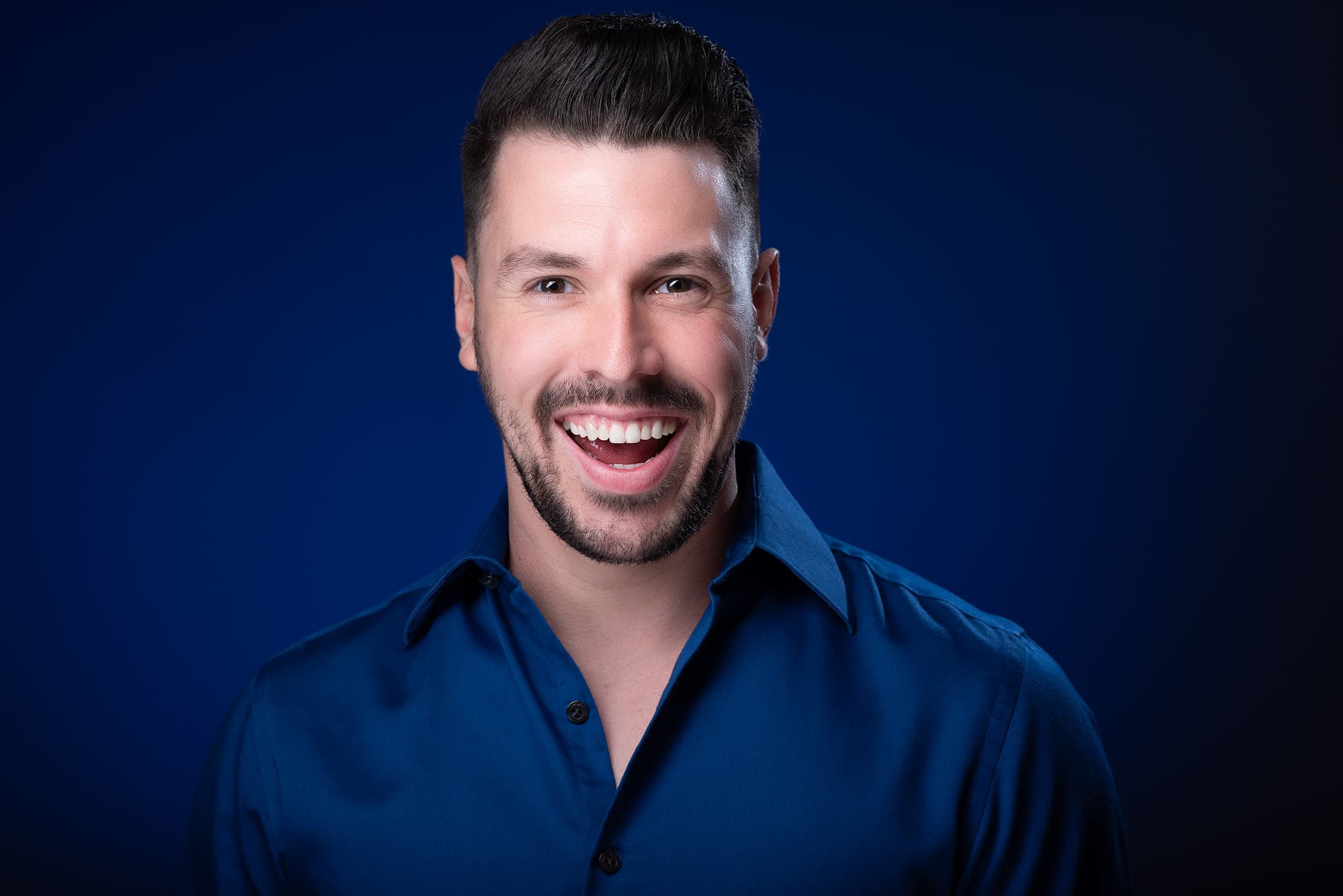 Atlanta actor headshot of Ethan Trace, dark-haired actor with a big genuine laugh in a navy shirt against a deep blue studio background