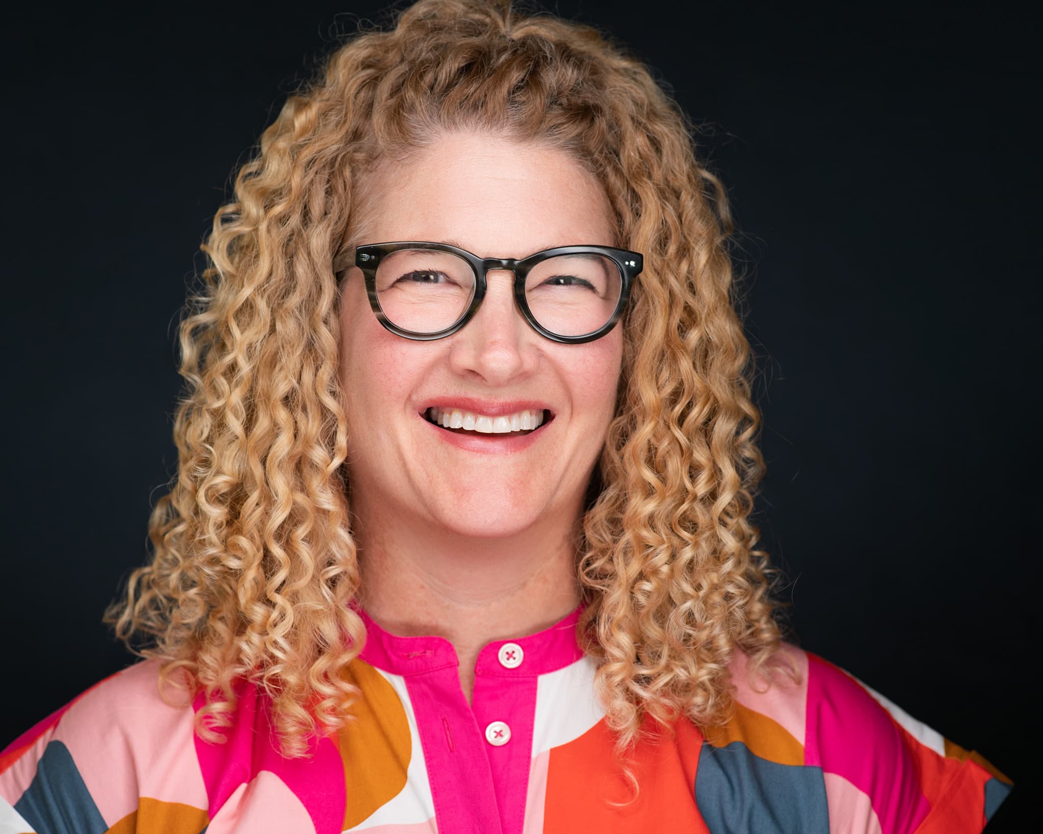 Professional woman's headshot with curly blonde hair, bold glasses and colorful pink blouse against black background