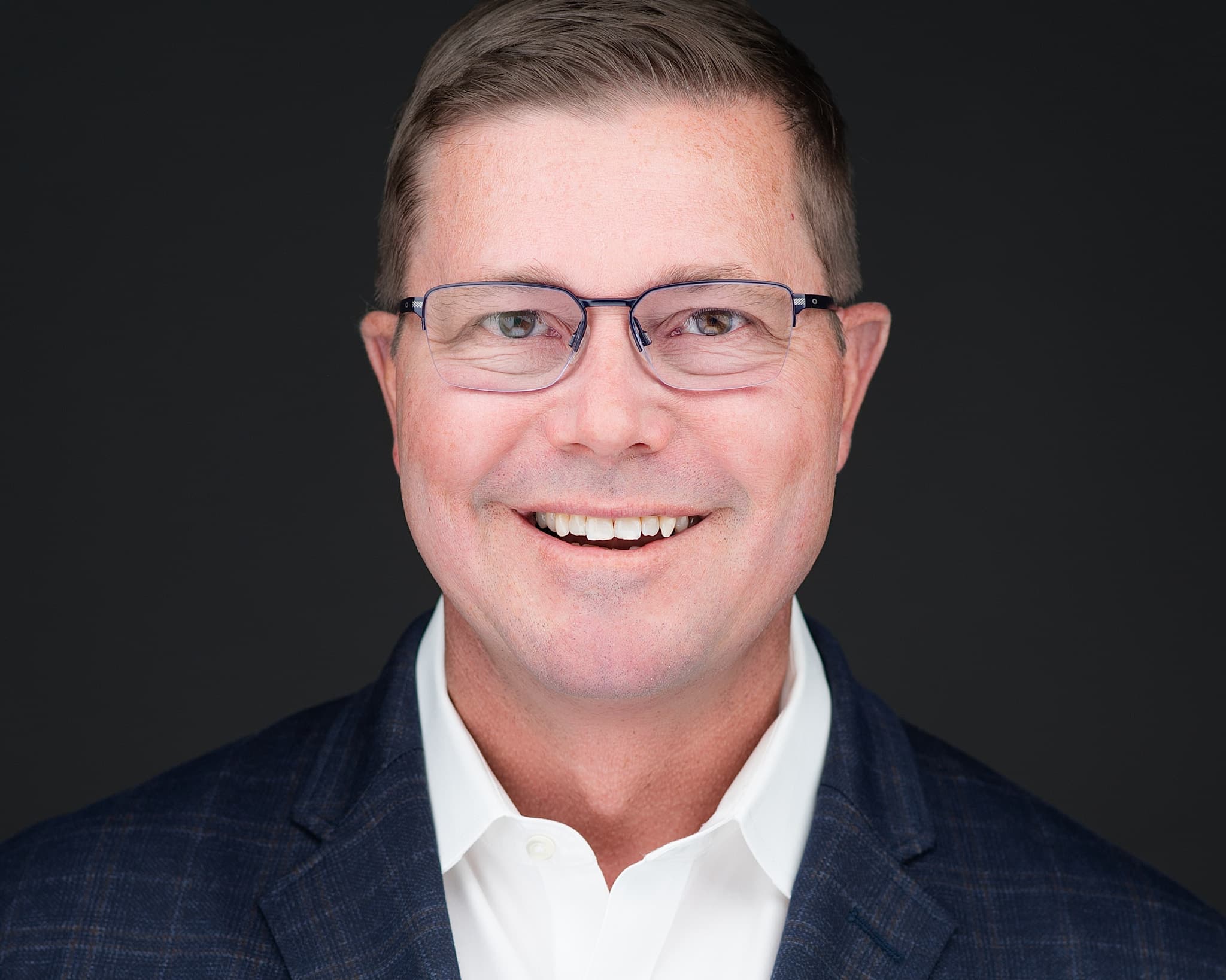 Male executive headshot with glasses, navy plaid blazer and white shirt, warm smile against dark background