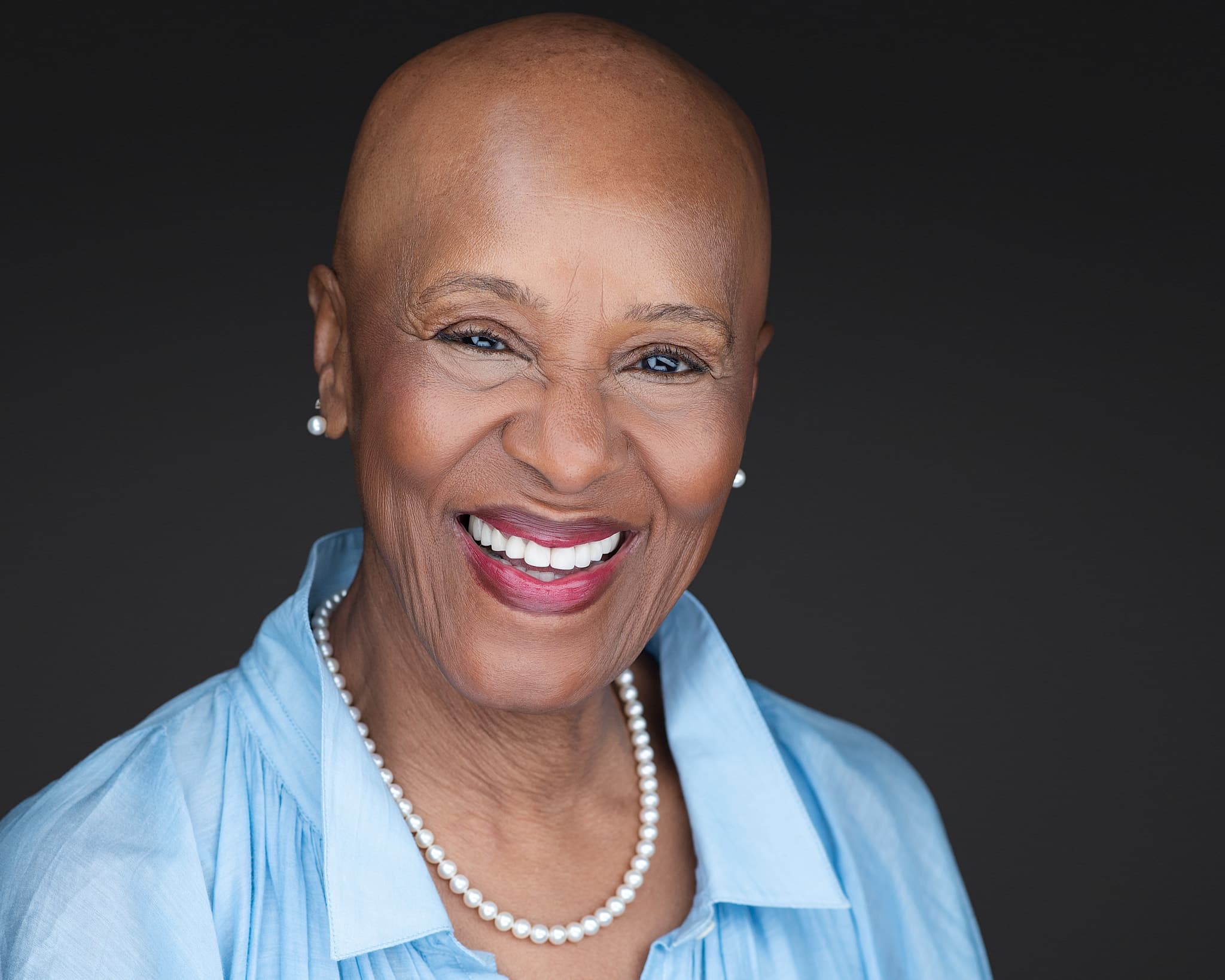 Executive woman's headshot with joyful smile, light blue blouse and pearls against dark background