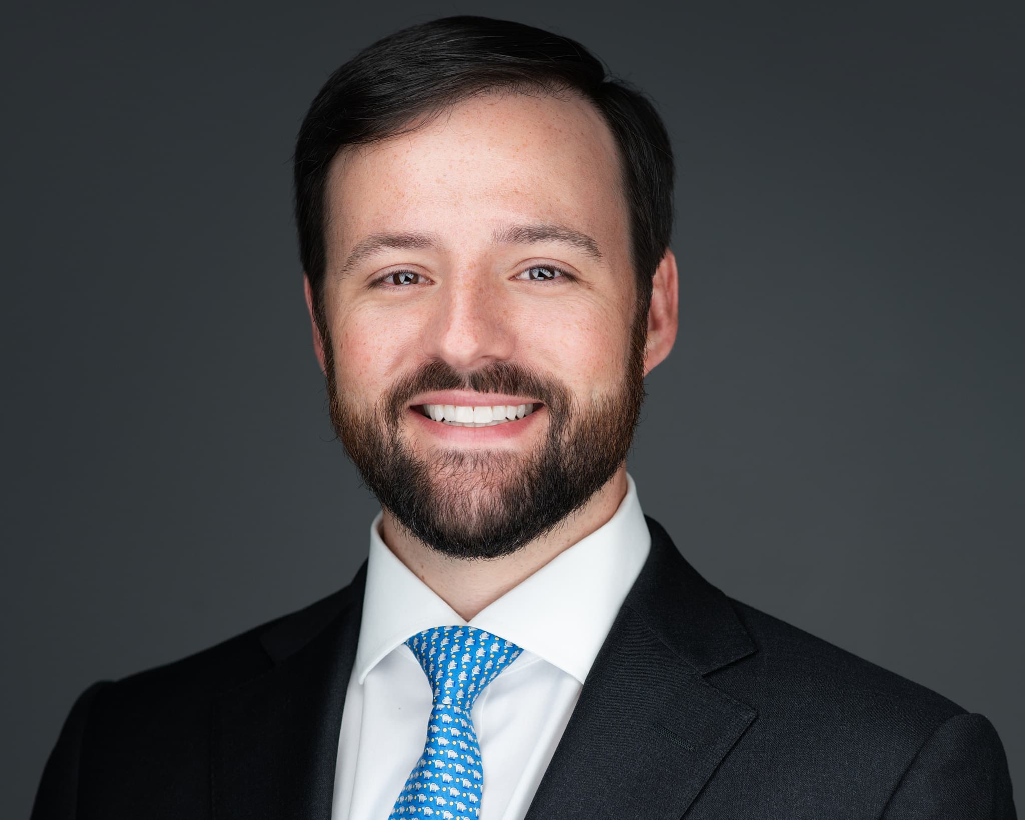 Male professional headshot with full beard, navy suit and light blue tie against dark gray background