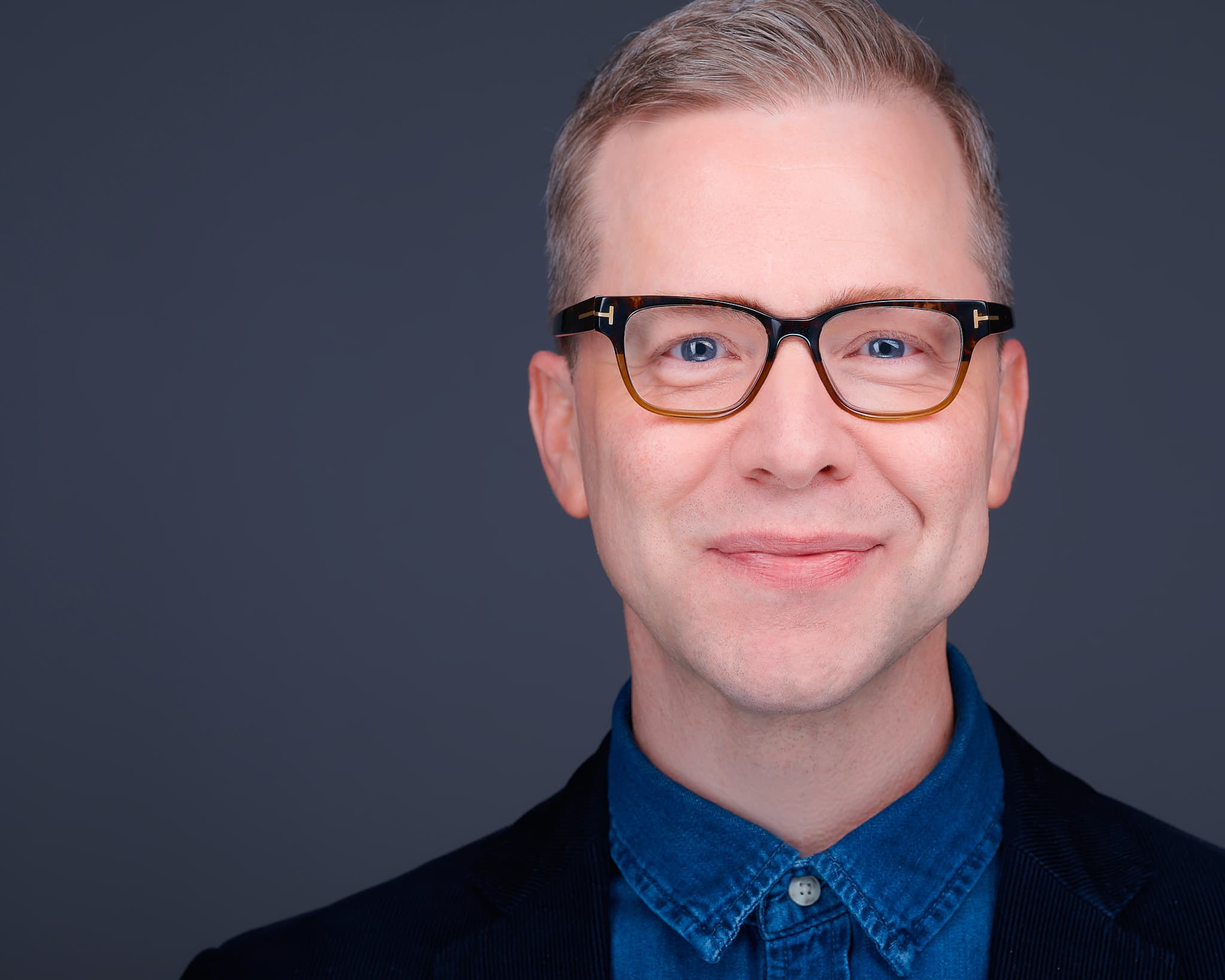 Corporate headshot of man in navy blazer and designer glasses against gray backdrop