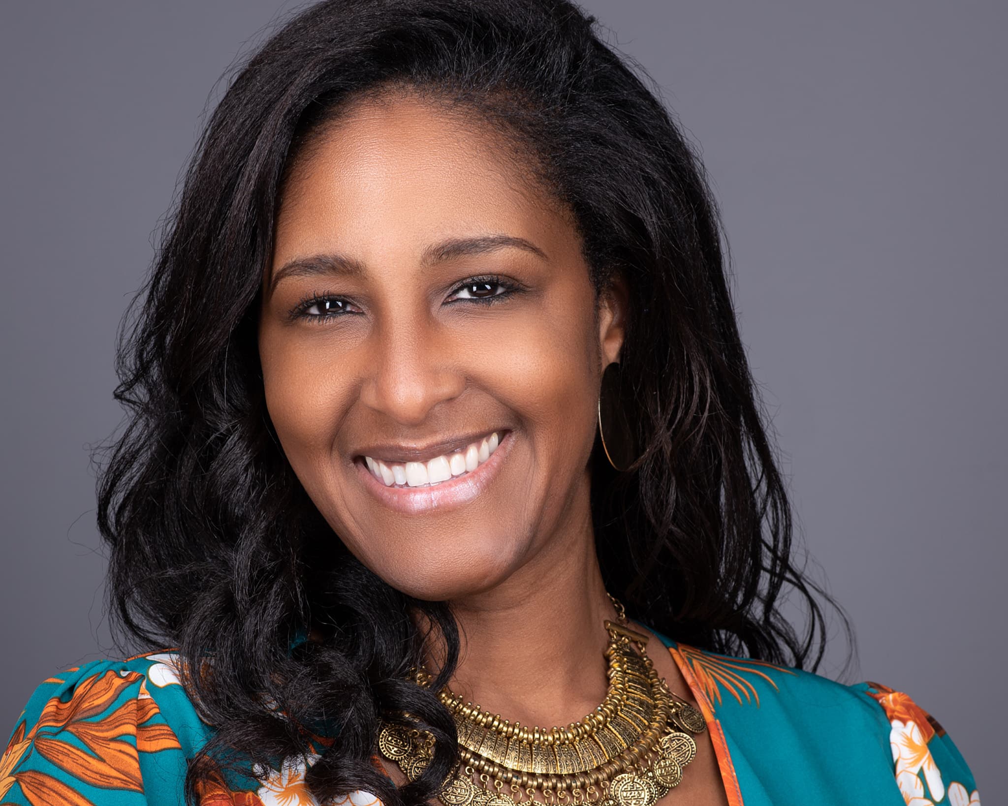 Female professional headshot with vibrant multicolored top and gold statement necklace on gray-blue background