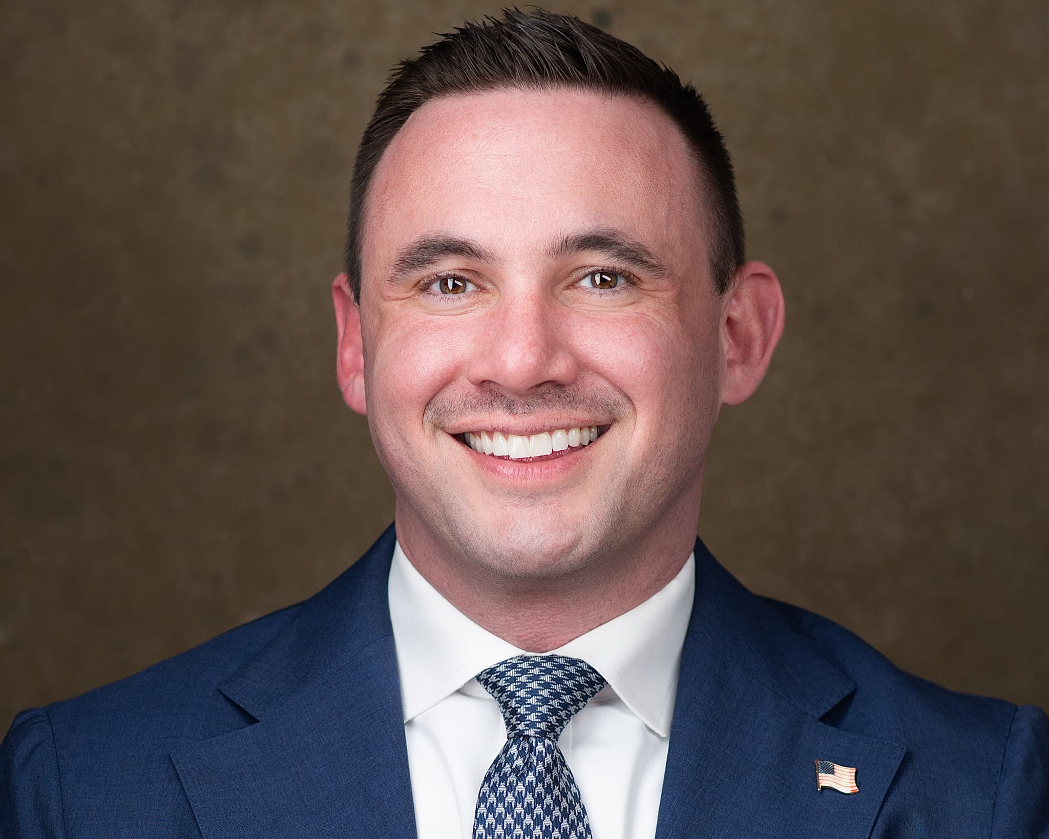 Male professional headshot in navy suit, patterned tie and lapel pin with confident smile against warm dark background