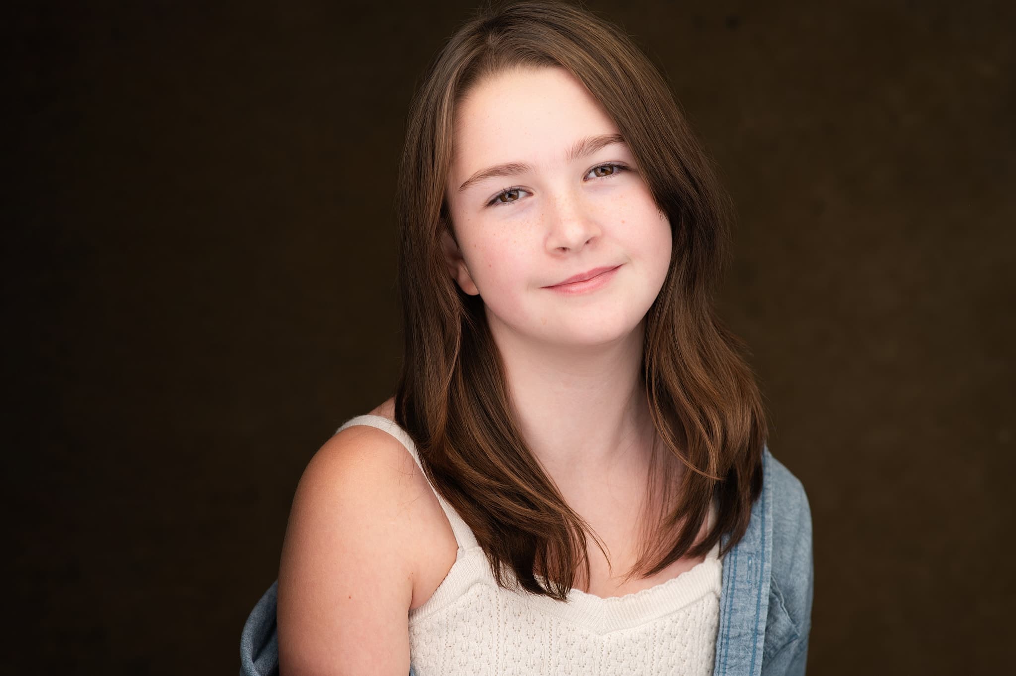 Atlanta tween actor headshot of Alisha Mayheu, young actress with long brown hair and a soft smile in a white top and denim shirt against a warm brown backdrop