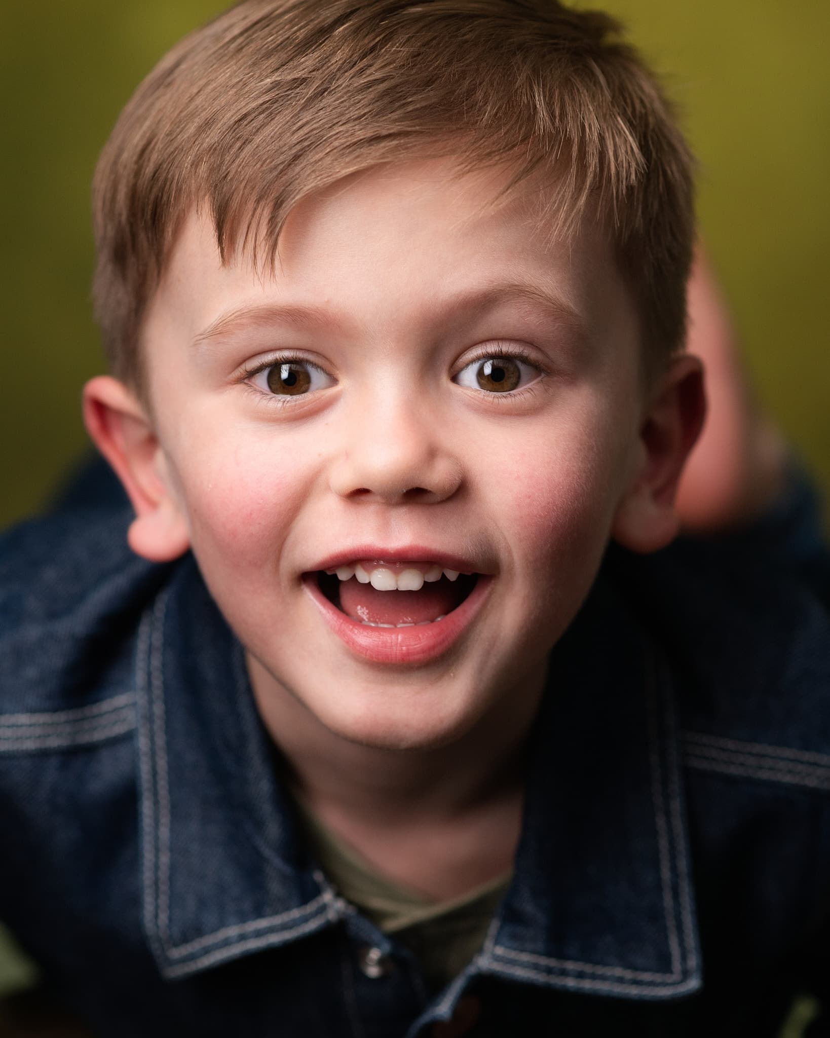 Atlanta child actor headshot of Griffin Guterman, young boy with sandy hair and a big playful grin in a denim jacket against an olive green backdrop