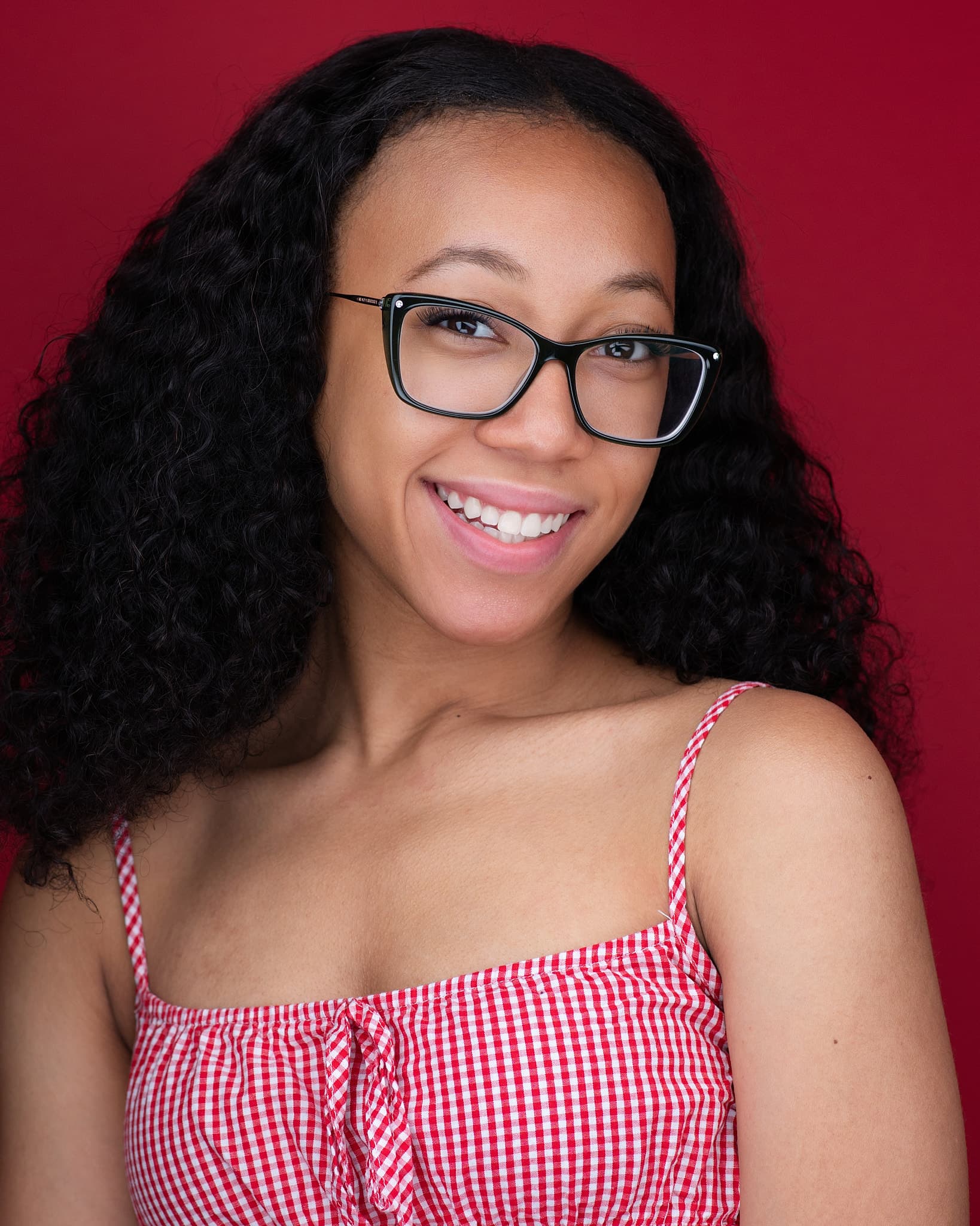 Atlanta teen actor headshot of a young performer with long dark curls and black glasses smiling in a red gingham top against a red backdrop