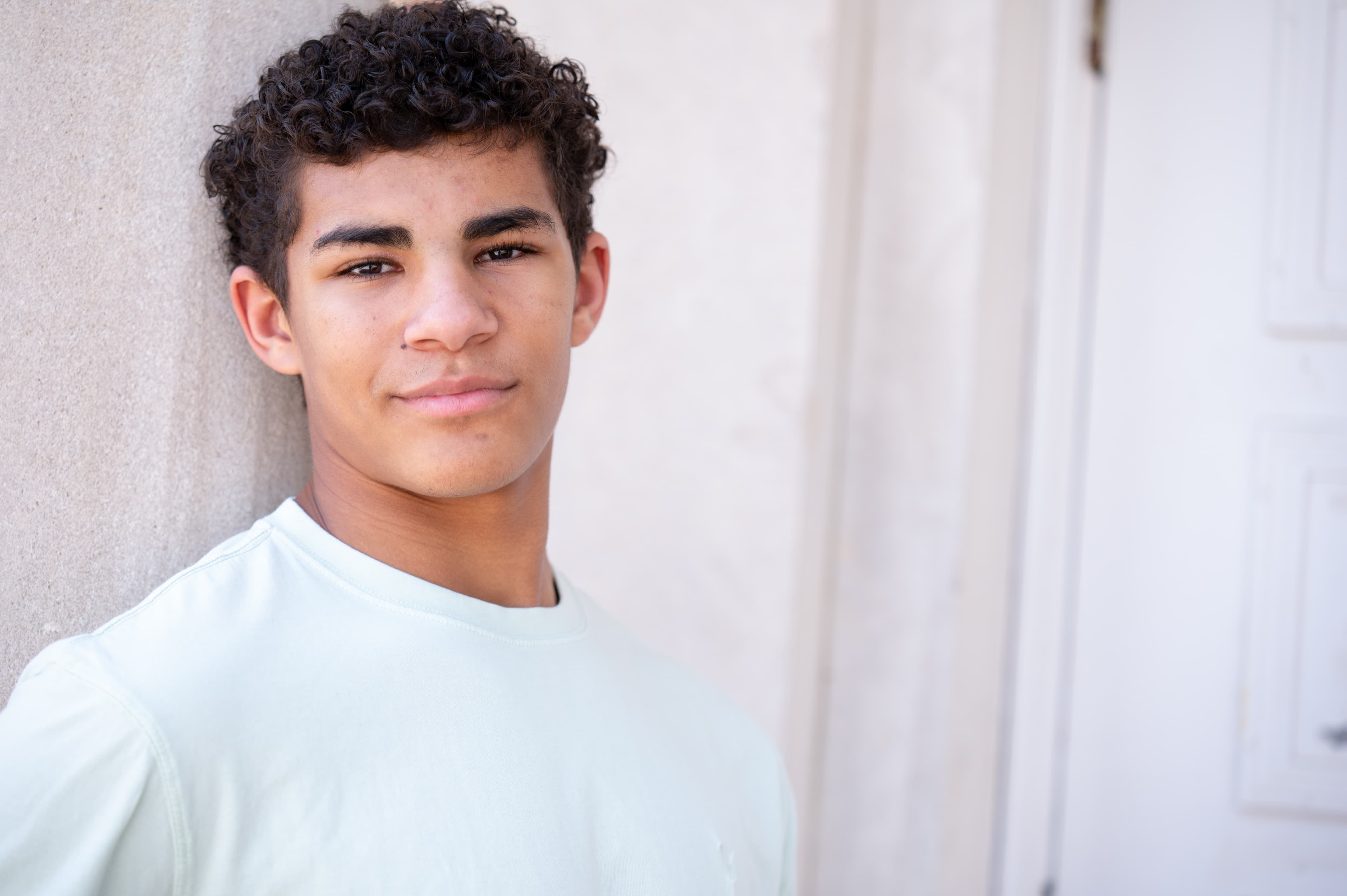 Atlanta commercial actor headshot of Grayson, young man in light blue tee with friendly approachable expression against neutral beige wall
