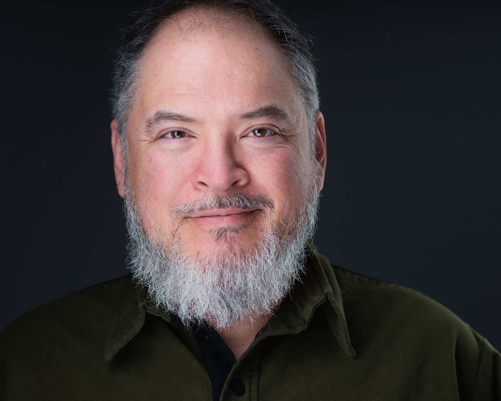 Male professional headshot with silver beard in olive shirt against moody dark background