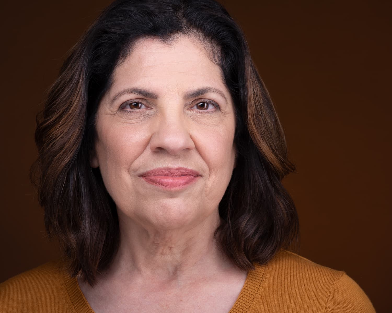 Atlanta theatrical actor headshot of Rose Bianco, mature woman in burnt orange top against warm brown background with contemplative gaze