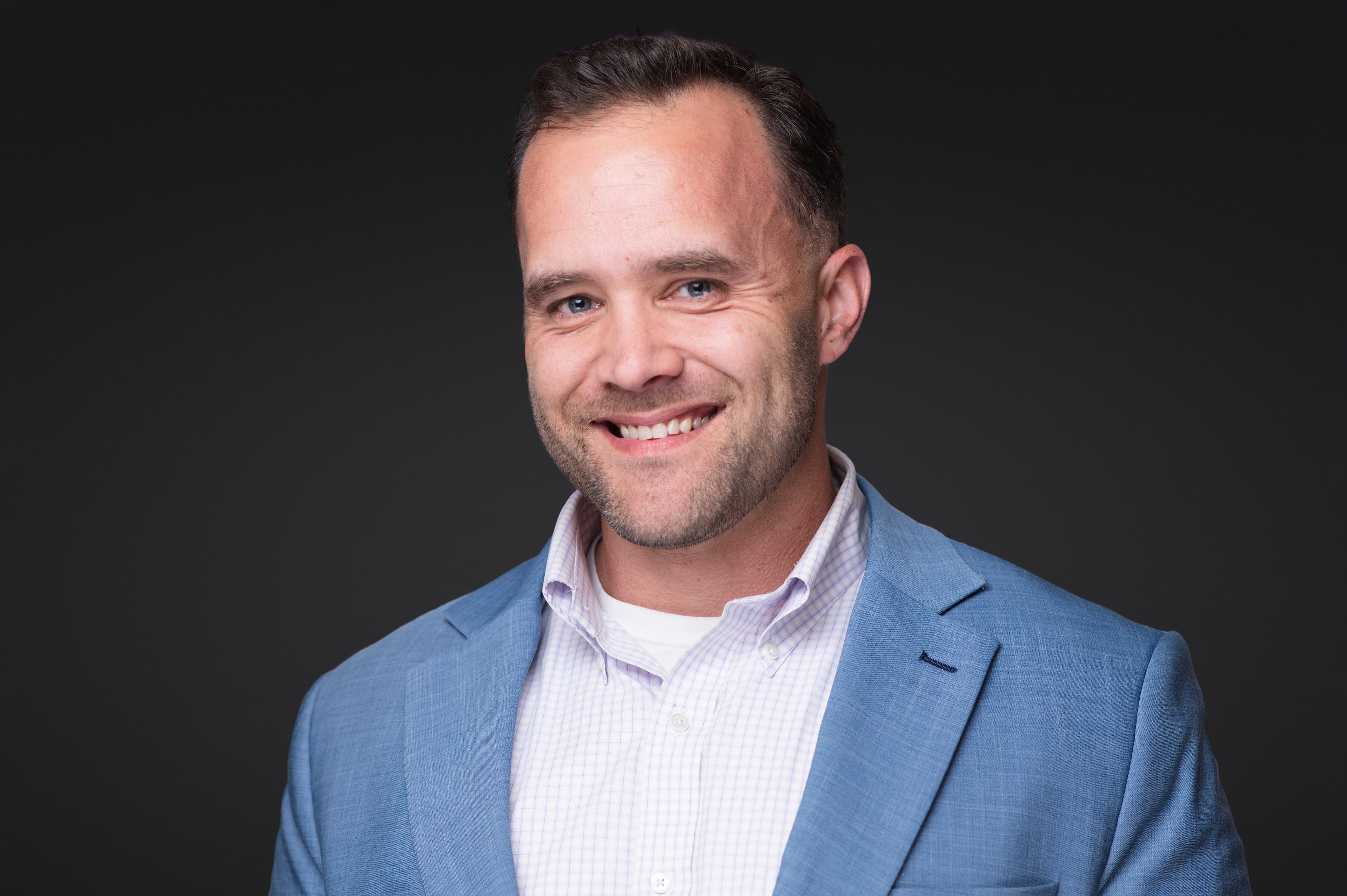 Professional headshot of man in blue blazer and striped pink shirt with beard and engaging warm smile