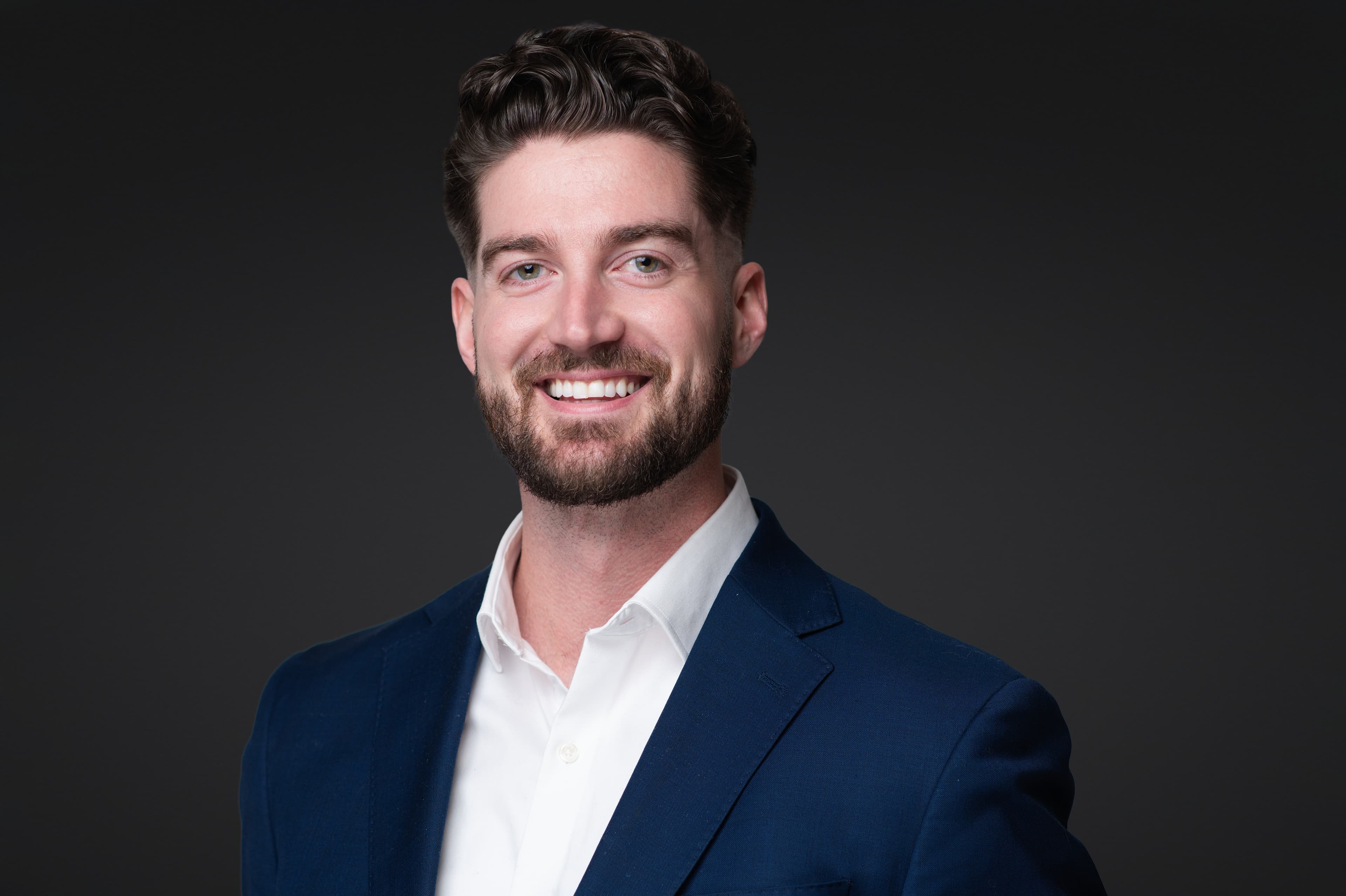 Young professional man corporate headshot with navy blazer, warm studio lighting, and confident smile against dark background