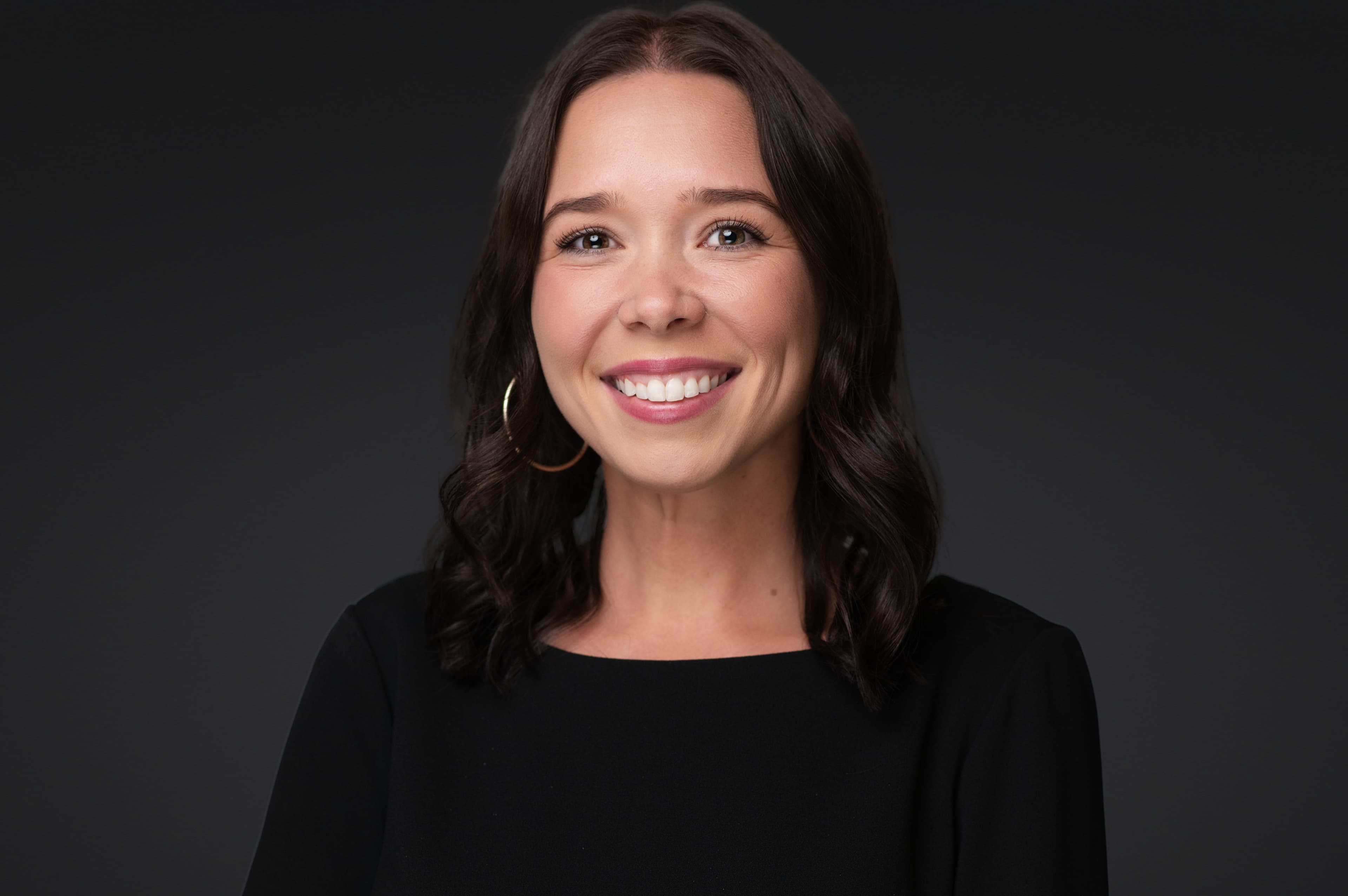 Professional woman headshot with long dark wavy hair, warm smile, and black top against neutral gray studio background