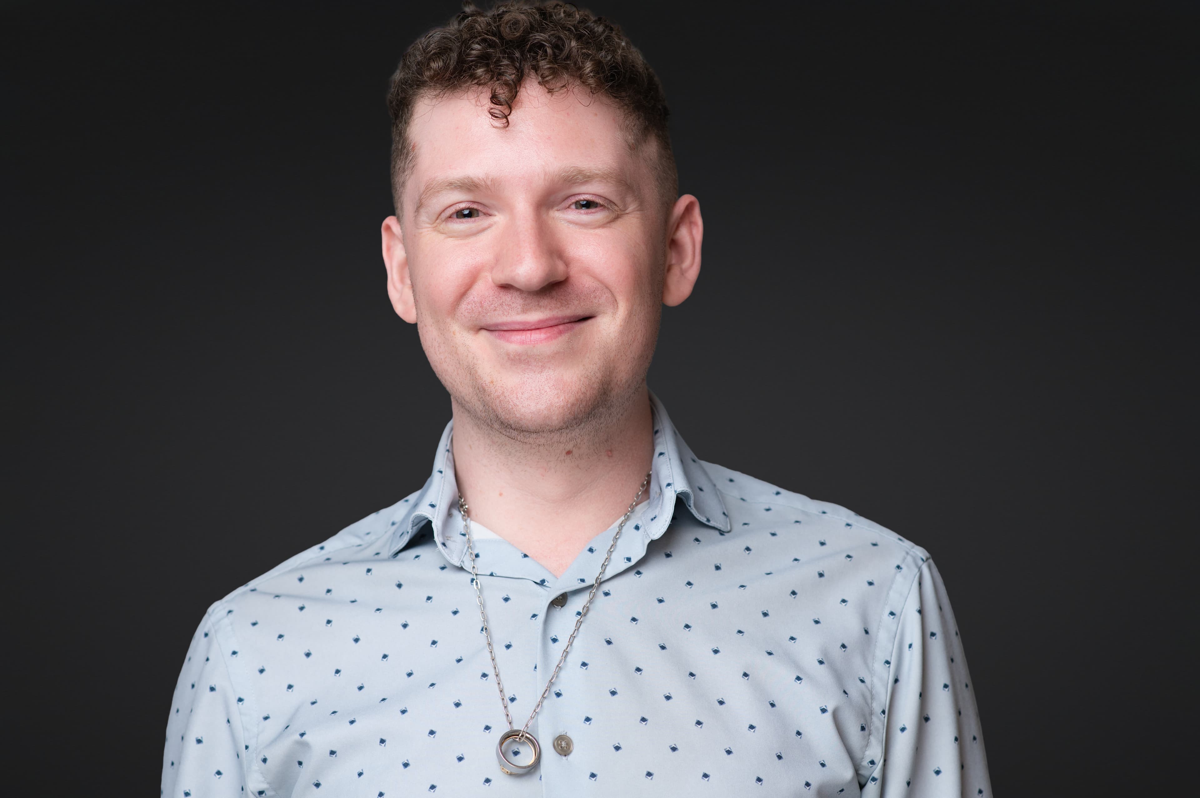Professional headshot of man in light blue patterned shirt with warm friendly smile and silver necklace