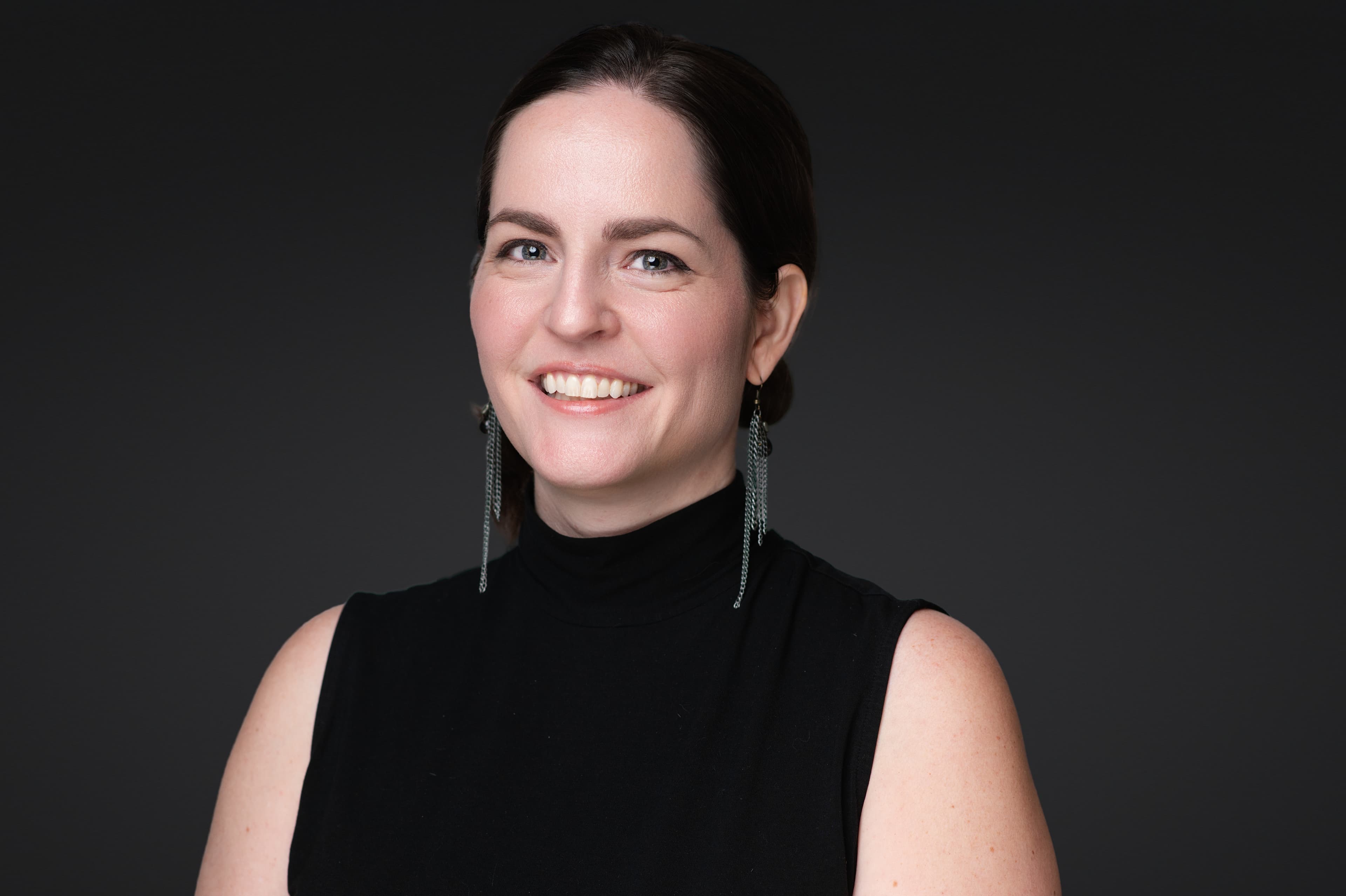 Professional headshot of woman in black sleeveless turtleneck with confident bright smile and silver earrings