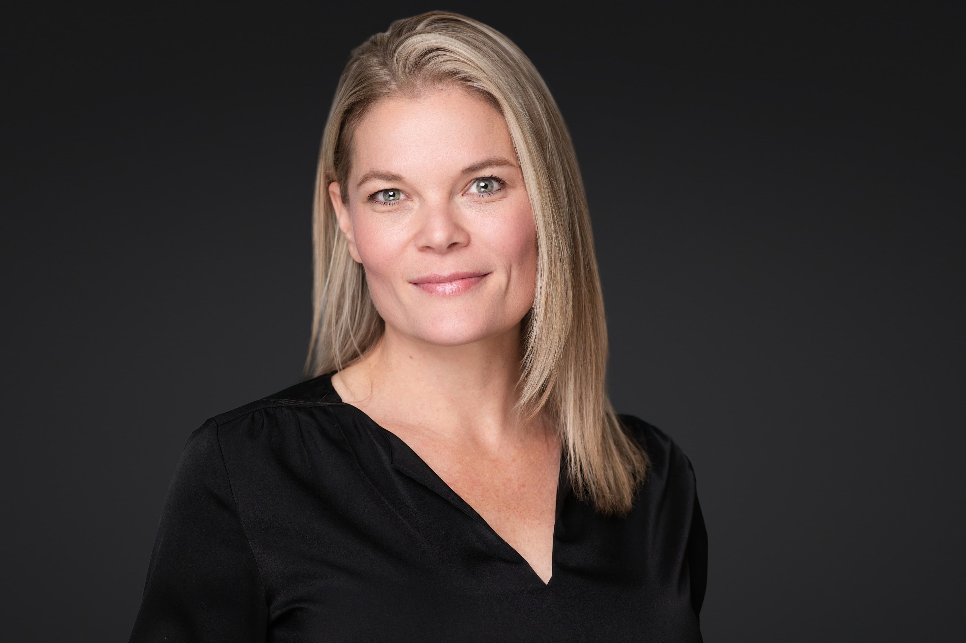 Mature professional woman headshot with blonde shoulder-length hair, composed expression, and black v-neck against dark background