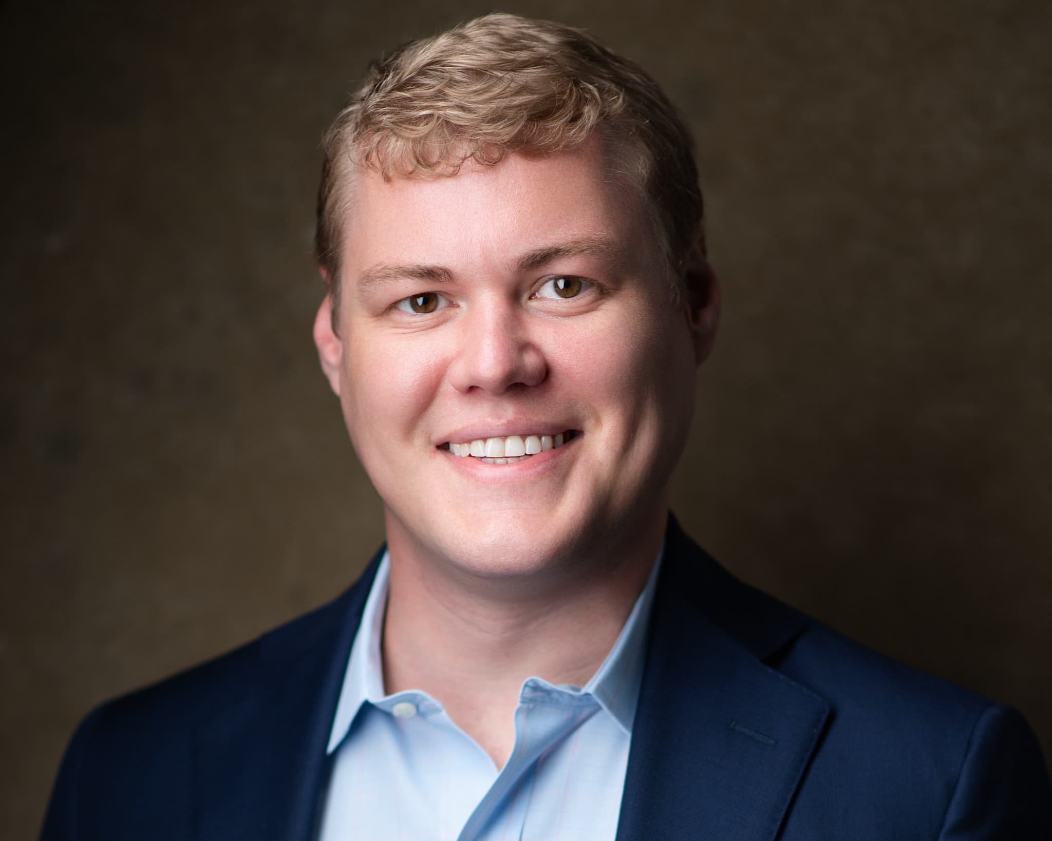 Male professional headshot in navy blazer and light blue shirt against warm brown backdrop
